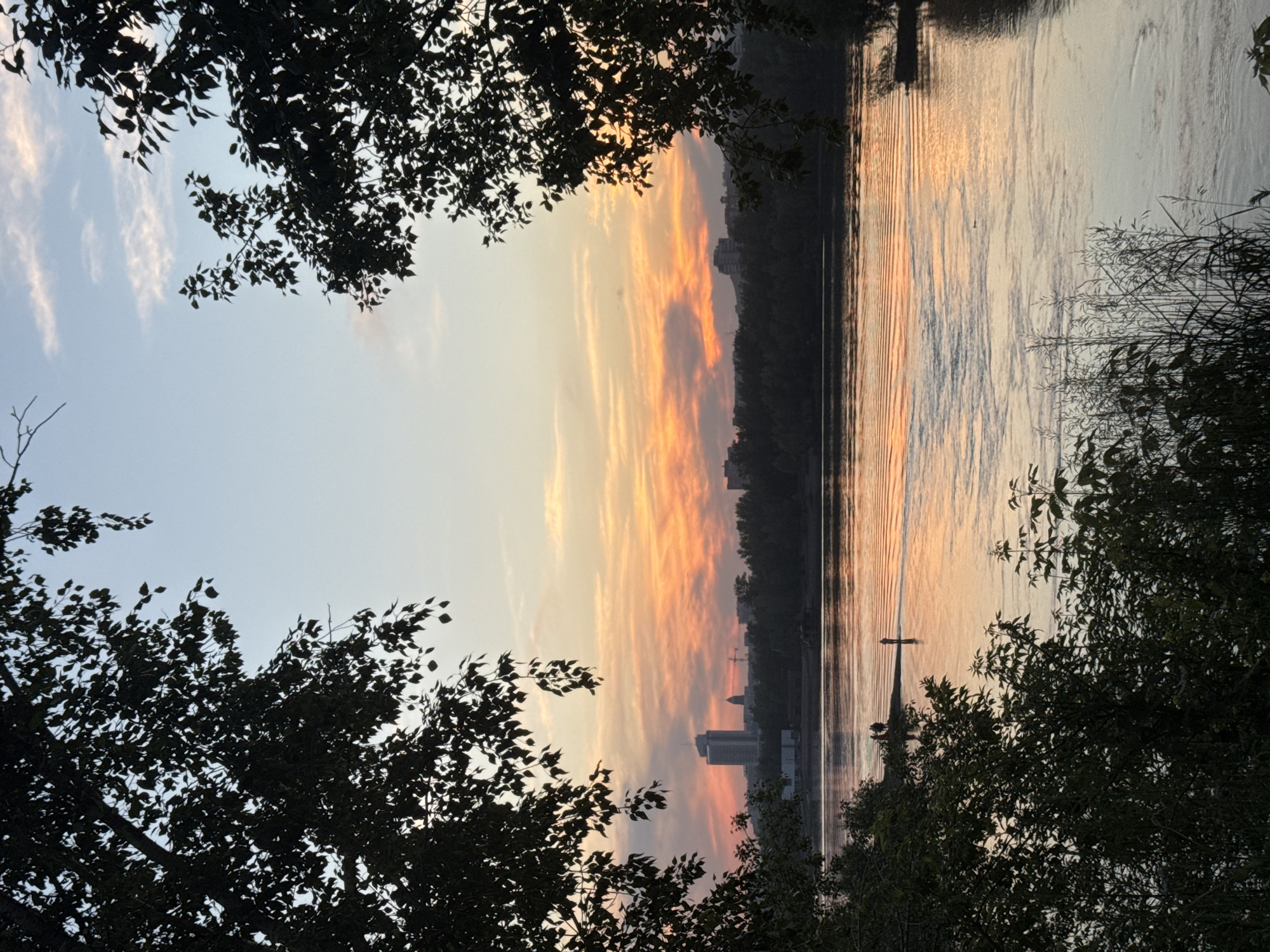 The image depicts a tranquil sunset scene over the Yenisei River in Krasnoyarsk, Russia. Silhouetted trees frame the left and right sides of the composition, their dark foliage contrasting with the vibrant sky. The sky transitions from a pale blue at the top to a fiery orange and pink hue near the horizon, with wispy clouds catching the last rays of the sun.
In the distance, a city skyline is visible, featuring buildings of various heights, including a prominent tall, slender building and a domed structure. The buildings are nestled amongst a line of dark trees that form the city's edge along the riverbank.
The Yenisei River dominates the middle ground, its surface reflecting the warm colors of the sunset, creating shimmering patterns of light. Small ripples indicate a gentle current. On a sandbar or shallow area extending into the river, several small human figures can be observed. One figure stands alone in the water, while a group of three is gathered on the edge of the sandbar. Their presence suggests a leisurely activity, possibly fishing or enjoying the view. The overall atmosphere is one of peacefulness and natural beauty at the end of the day.
