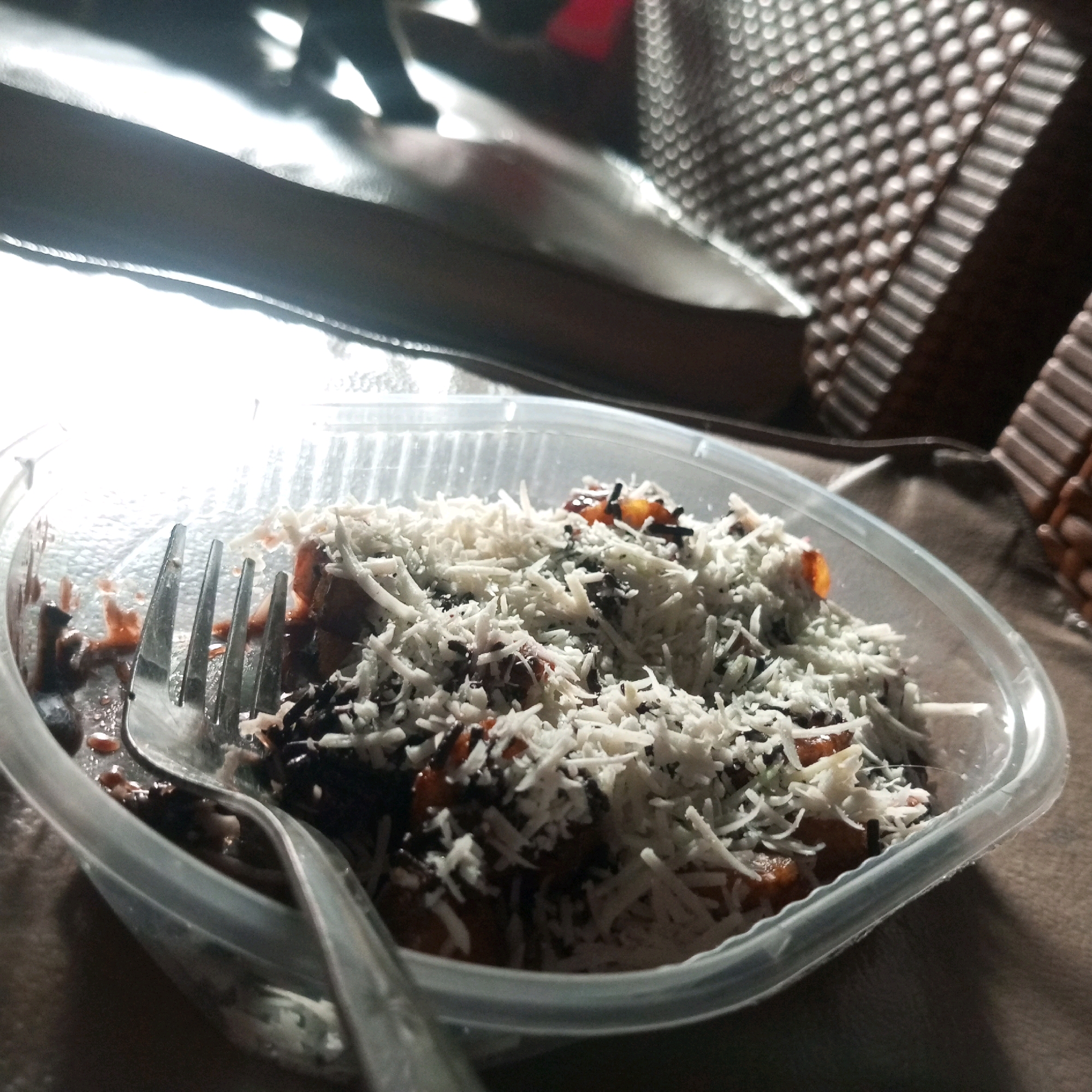 A close-up, low-angle shot captures a clear plastic container filled with a dessert, likely chocolate and cheese flavored, placed on a brown surface. A silver fork rests on top of the dessert, partially submerged. The background is blurred, with hints of a patterned surface and a dark, reflective area suggesting furniture. The lighting is dim, with harsh highlights, indicating an indoor setting, possibly a cafe or dining area in North Jakarta, Indonesia.