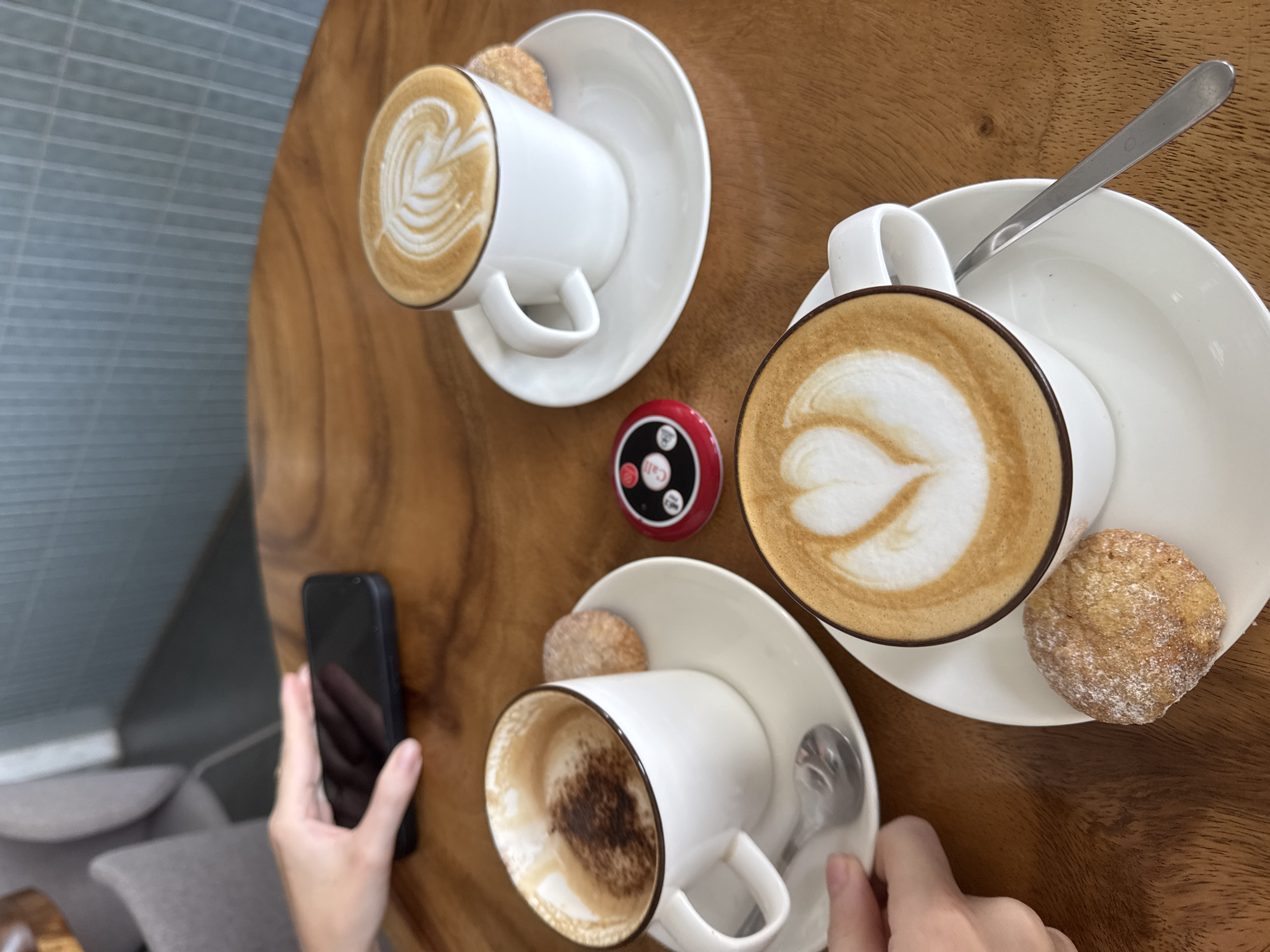 The scene captures a moment at a cafe in an unspecified city. Three white coffee cups with latte art designs sit on matching saucers on a wooden table. One cup is almost empty, with remnants of coffee and cocoa powder visible inside. Each saucer holds a small, round cookie dusted with powdered sugar. A metal spoon rests on one of the saucers. Between the cups, a small, red call button is placed on the table. A person's hands are visible, one holding a black smartphone and the other steadying a cup. In the background, a glimpse of a gray upholstered chair and a textured wall with horizontal lines is visible.
