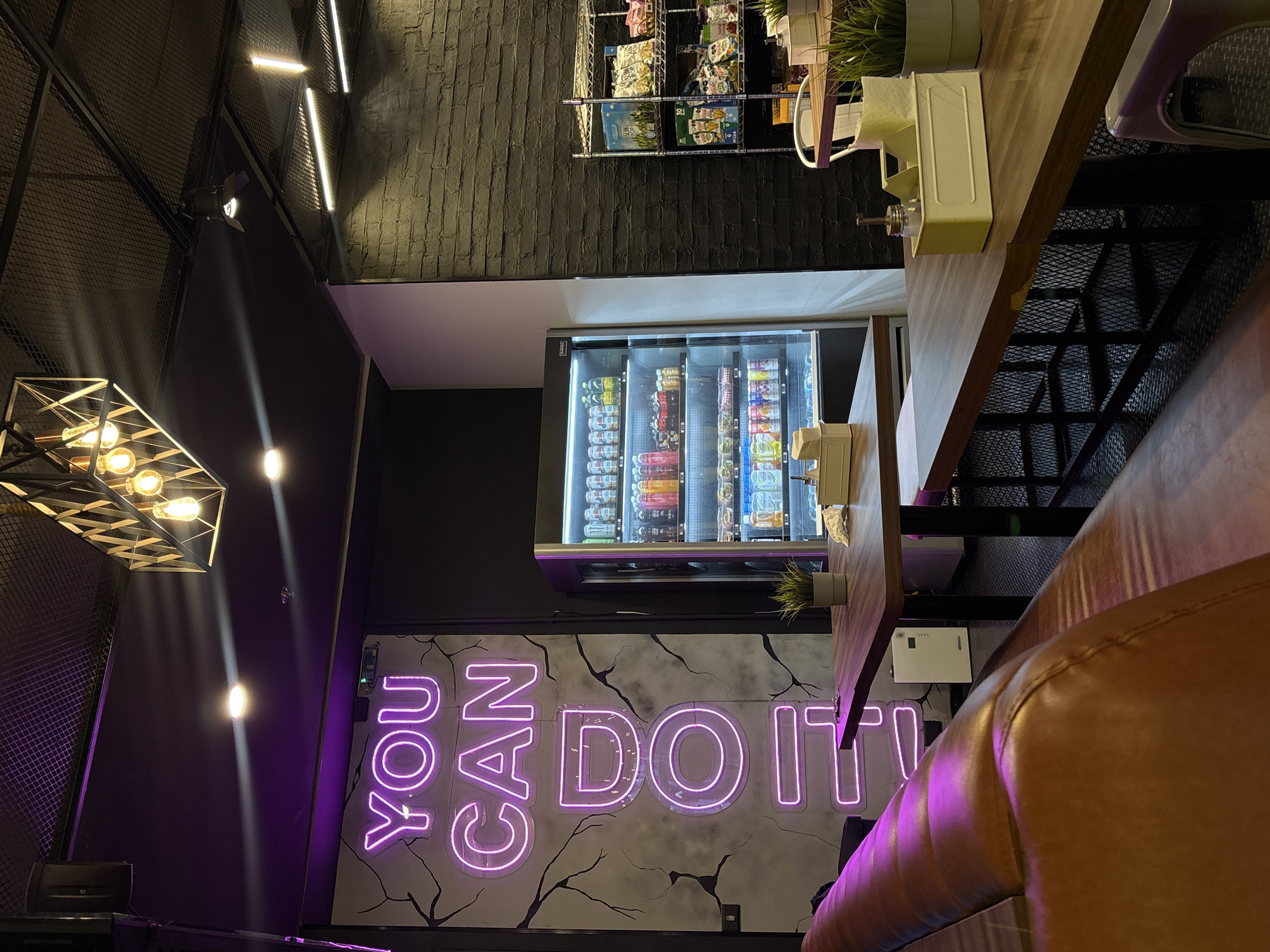 The image shows the interior of a business in San Bernardino Tlaxcalancingo, Mexico. A neon sign reading "YOU CAN DO IT" is mounted on the wall with a faux cracked-paint effect. Above it is a glass-fronted refrigerated vending machine stocked with beverages and snacks. A shelving unit displaying bagged snacks is affixed to a brick wall. A light fixture casts an indirect light on the left side of the image. Tables are placed along the wall, with leather seating on the right side of the image.