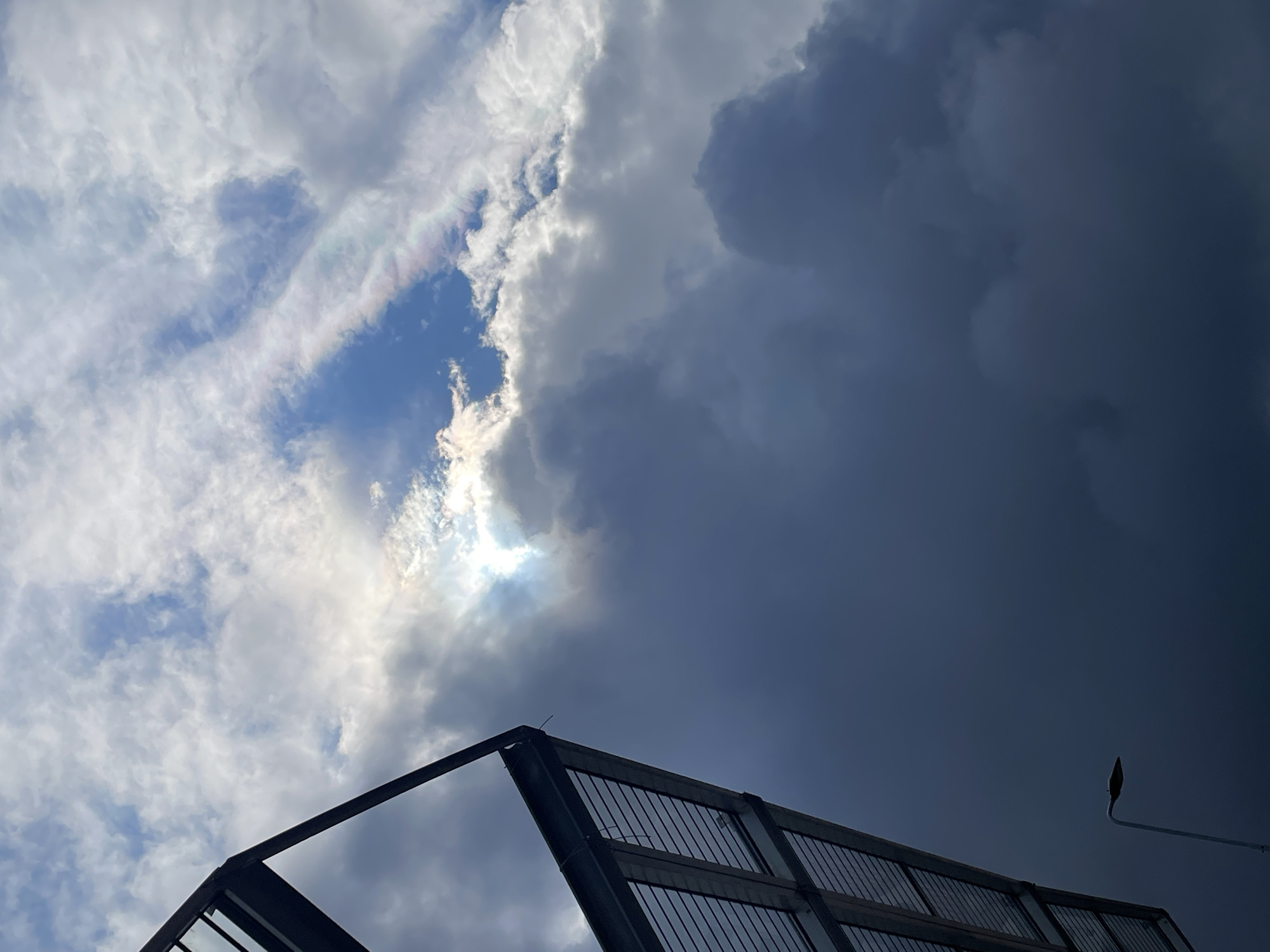 The image shows a view of the sky with a mix of light and dark clouds. A bright opening reveals a patch of blue sky with the sun shining through. Below the clouds, a portion of a metallic structure, possibly a roof or part of a building, is visible. A streetlight is seen to the right of the frame. The location appears to be in an urban setting, as indicated by the presence of the structure and streetlight, but the specific city is unknown.