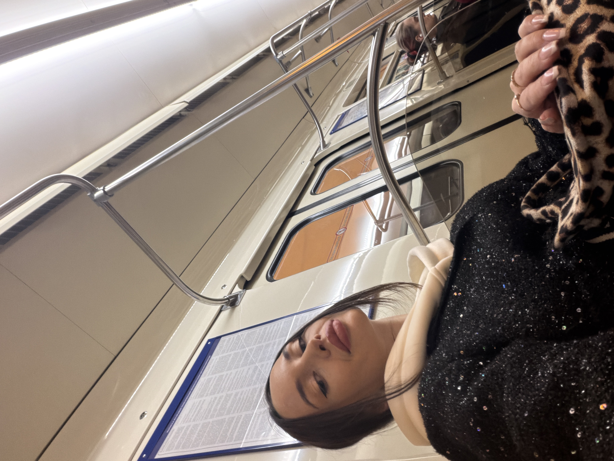 A woman with dark hair and heavy eye makeup is the focal point of the photograph, taken at an unusual angle. She is wearing a black, possibly sequined top over a light-colored hoodie, and holding a leopard-print accessory. The background suggests the interior of a subway car, likely in Saint Petersburg, Russia, recognizable by the typical train design, including metal railings, interior panels, and windows. A blue information panel with text is visible on the wall. The overhead lights are on, providing illumination within the subway car.