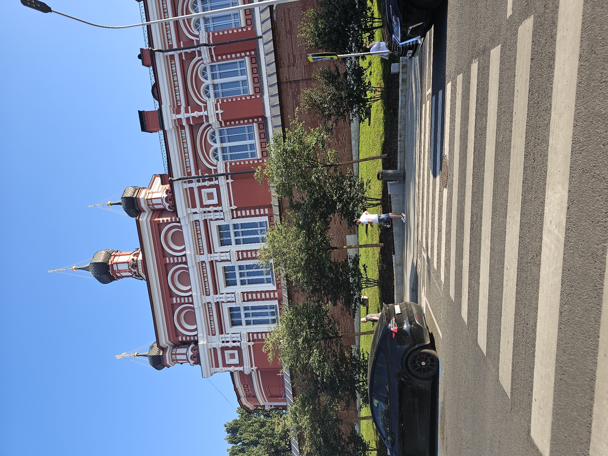 The image shows a wide shot of a street in Moscow, Russia, with a prominent red and white building in the background, characteristic of Russian Orthodox architecture with its onion domes and ornate facade. The building is situated behind a red brick wall and a line of trees. In the foreground, a pedestrian crossing is visible on an asphalt road. Two black cars are present; one is parked near the curb on the left side of the frame, and the front of another car is visible on the right side, also near the pedestrian crossing. Two individuals are seen standing near the curb between the trees and the building, and another person is visible on the far right near the second car. The sky is clear and blue, indicating fair weather. The scene appears to be a public urban space during daylight hours.