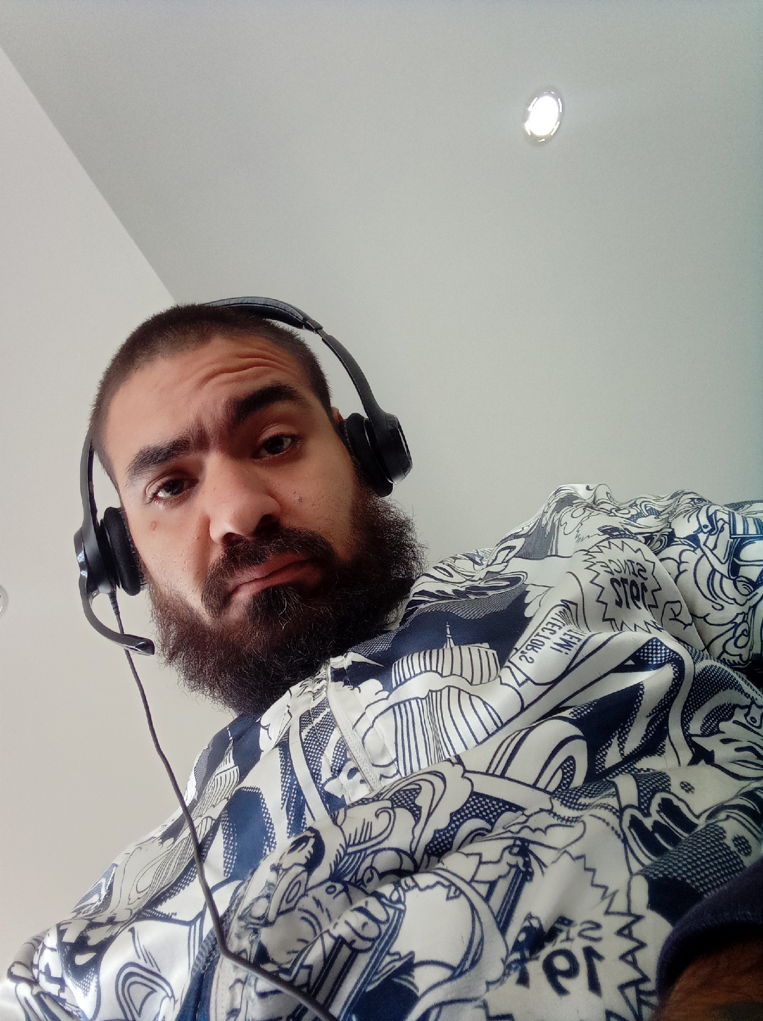 A man with dark hair and a beard is wearing a headset and a white shirt with a blue graphic pattern. The photo is taken from a low angle looking up at the man. A light fixture is visible on the white ceiling. The location, Parroquia Guatire, Venezuela, cannot be determined from the provided image.