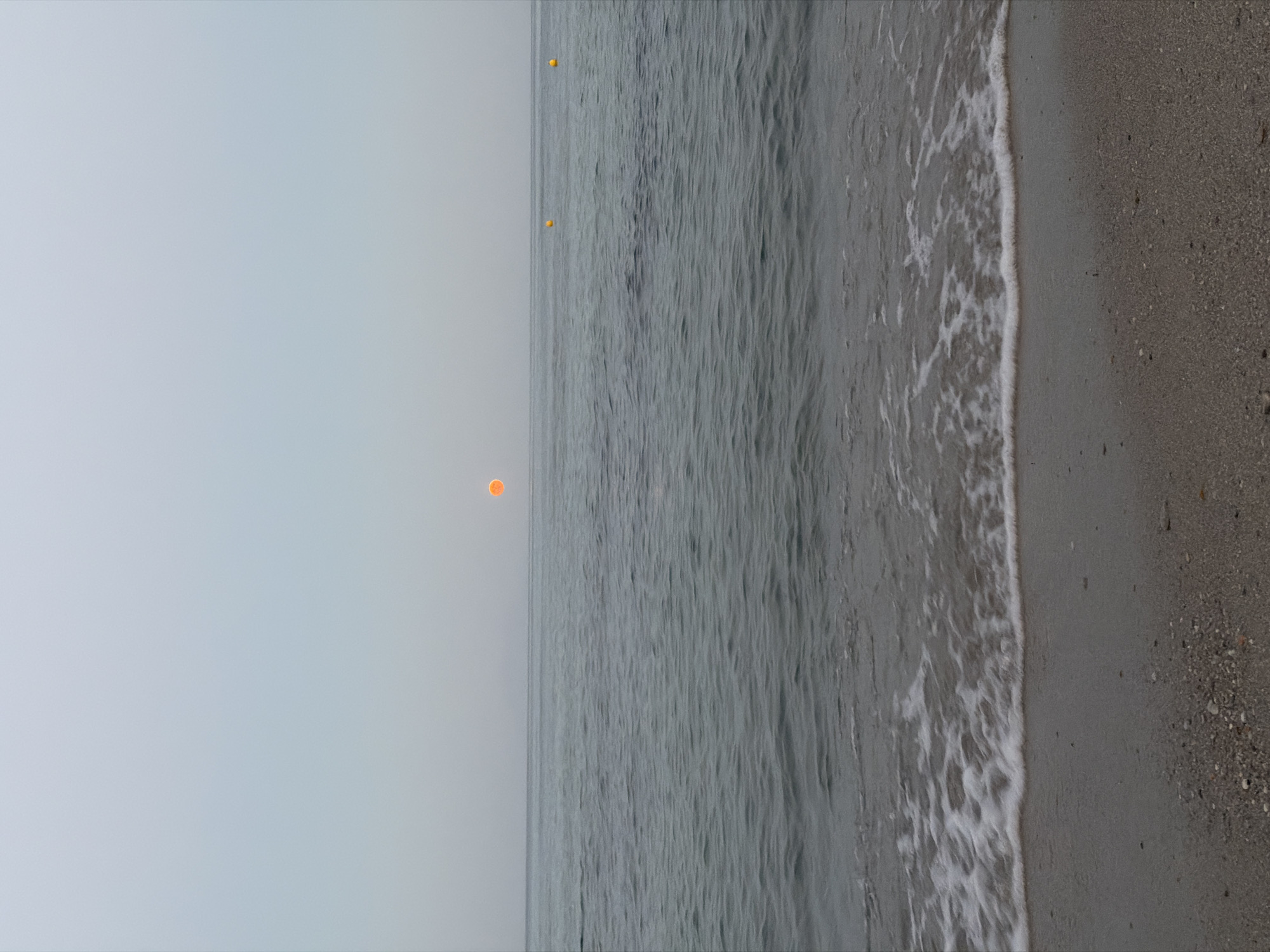 A wide shot of a calm sea under a hazy sky shows a single orange sun on the horizon. The sun is located slightly left of the center of the image. The water is a muted gray-blue with small ripples. A gentle wave is breaking on a sandy beach in the foreground. The sand is a light brown color and appears to be mixed with small pebbles and shells. Two yellow buoys are visible in the distance, one to the left of the sun and another further to the right. The overall atmosphere is serene and muted, typical of a cloudy or misty day. The location is Golden Beach, Greece.