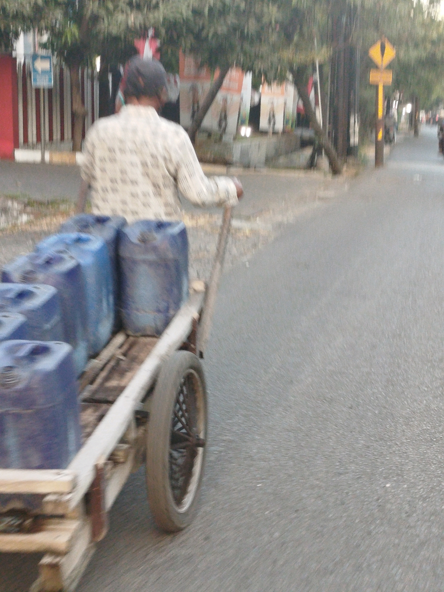 A person is pushing a cart loaded with several blue plastic containers down a street in Surabaya, Indonesia. The cart is made of wood and has one visible wheel. The person is wearing a light-colored patterned shirt and a dark cap. The street appears to be paved, with trees lining the side. There are buildings and signs visible in the background, suggesting an urban environment. The image is blurred, indicating movement.
