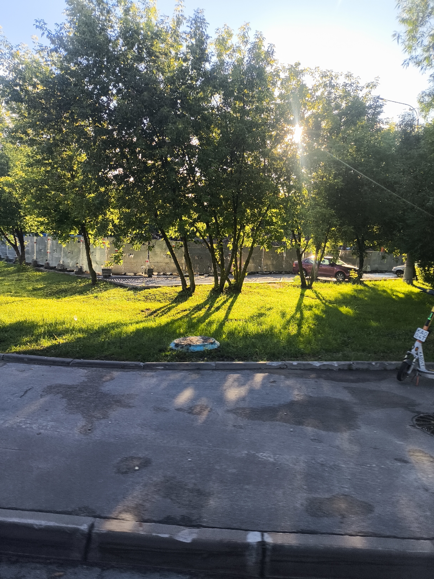 A sun-drenched scene depicts a suburban or urban environment with trees casting long shadows on a lush green lawn. The sun's rays are visible through the leaves, creating a bright glare in the upper right portion of the image. A red car is parked on the street in the background, partially obscured by trees. A white electric scooter with an orange handlebar is parked on the right side of the frame, facing left. The foreground shows a wet asphalt road with a concrete curb. A circular manhole cover with a blue rim is visible on the grass. In the far background, a metallic fence with reflective panels is visible. The sky is clear and blue.