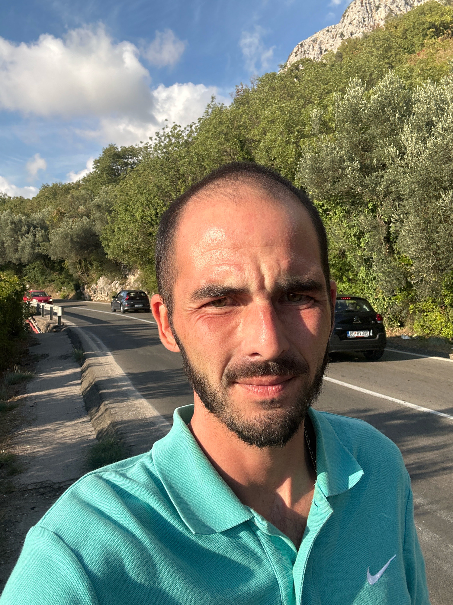A man with a beard and short hair, wearing a teal polo shirt, is taking a selfie outdoors. He is standing on the side of a road that curves to the left. Behind him, the road continues and is bordered by lush green trees on the right. In the distance, a black car is visible on the road. To the left of the road, there is a concrete barrier. The background features a steep, rocky mountain covered in trees, and a bright blue sky with scattered white clouds. The scene appears to be in the city of Drobnići, Montenegro.
