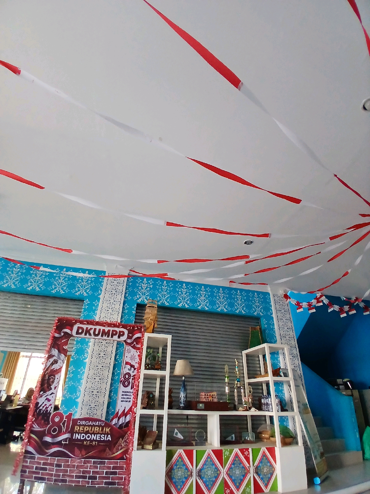 The image depicts an indoor scene, likely a decorated space within the city of Tarempa Timur, Indonesia. Red and white streamers, reminiscent of the Indonesian flag, are suspended from the ceiling, creating a festive atmosphere. The walls are painted blue with a white ornate stencil pattern.
In the foreground, a large, red and white photo booth frame is set up, featuring text that reads "DKUMPP" and "DIRGAHAYU REPUBLIK INDONESIA KE-81," celebrating the 81st anniversary of the Republic of Indonesia. Behind the photo booth, a tall, white shelving unit displays various items, including trophies, a lamp, decorative vases, and what appear to be framed certificates or awards.
To the left of the photo booth, a window allows natural light to enter the space, and a person can be seen working at a desk in the background. To the right of the shelving unit, a blue staircase ascends, with a portion of a blue wall and more red and white flag-like decorations visible along the railing. The floor area in front of the shelving unit is decorated with colorful geometric patterned panels. The overall impression is one of a celebratory event or commemoration.