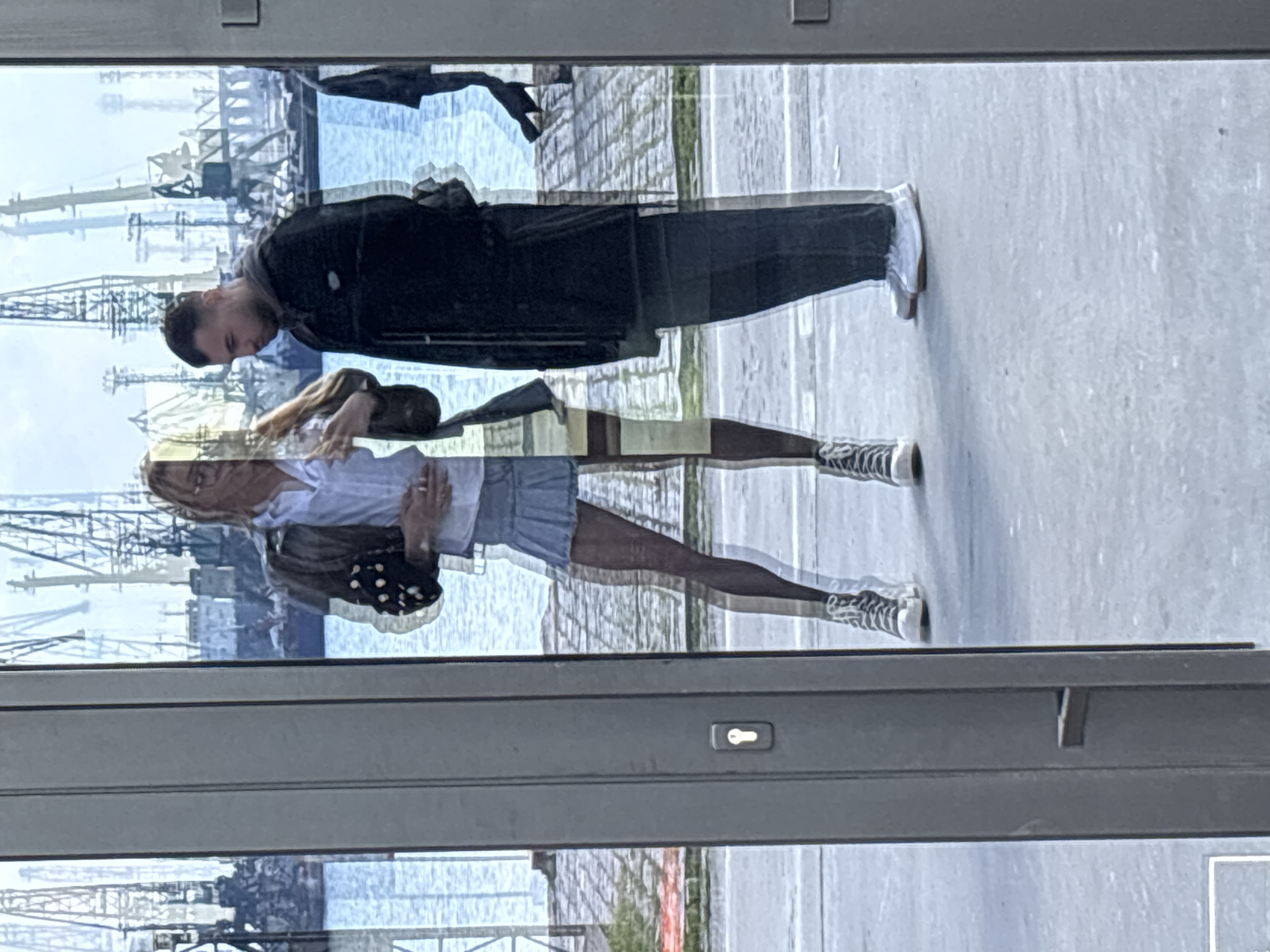 The image is a reflection in a window. Reflected are a man and a woman, superimposed over a cityscape. The man is wearing a dark jacket and dark pants, and white shoes. The woman has blond hair and is wearing a white shirt, jean skirt, and sneakers. She is also carrying a purse. The background appears to be a waterfront scene with cranes, suggesting a port or harbor area. Given the description, the scene is likely in Varna, Bulgaria, potentially near the Varna Port or along the coast. The window also reflects the building interior.