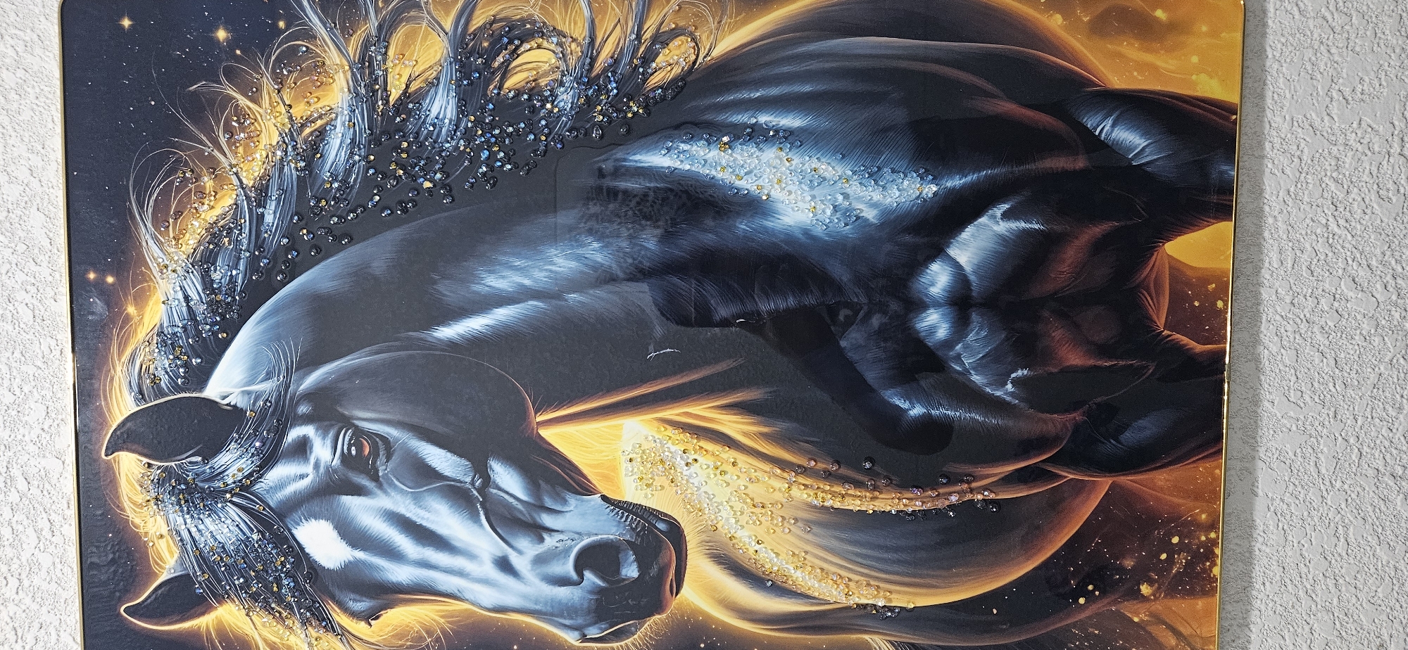 This is a close-up, portrait-style image of a black horse, rendered in a painterly, digital art style. The horse is depicted from the chest up, facing slightly to the left. Its mane and forelock are flowing dynamically and appear to be adorned with numerous small, sparkling embellishments that resemble crystals or glitter, catching the light. The background is a dark, starry sky with vibrant, glowing orange and yellow light emanating from behind the horse, suggesting a fiery or celestial theme. There are also flecks of similar glowing particles scattered throughout the background and on the horse's body, particularly on its chest and along the mane. The horse itself has a glossy, almost metallic sheen, with sculpted musculature clearly defined. A white blaze marks its forehead. The image is framed by a thin, gold-colored border, and the textured surface of a wall is visible below the artwork. The image does not contain any discernible elements that would allow for its location to be identified as Dushanbe, Tajikistan.