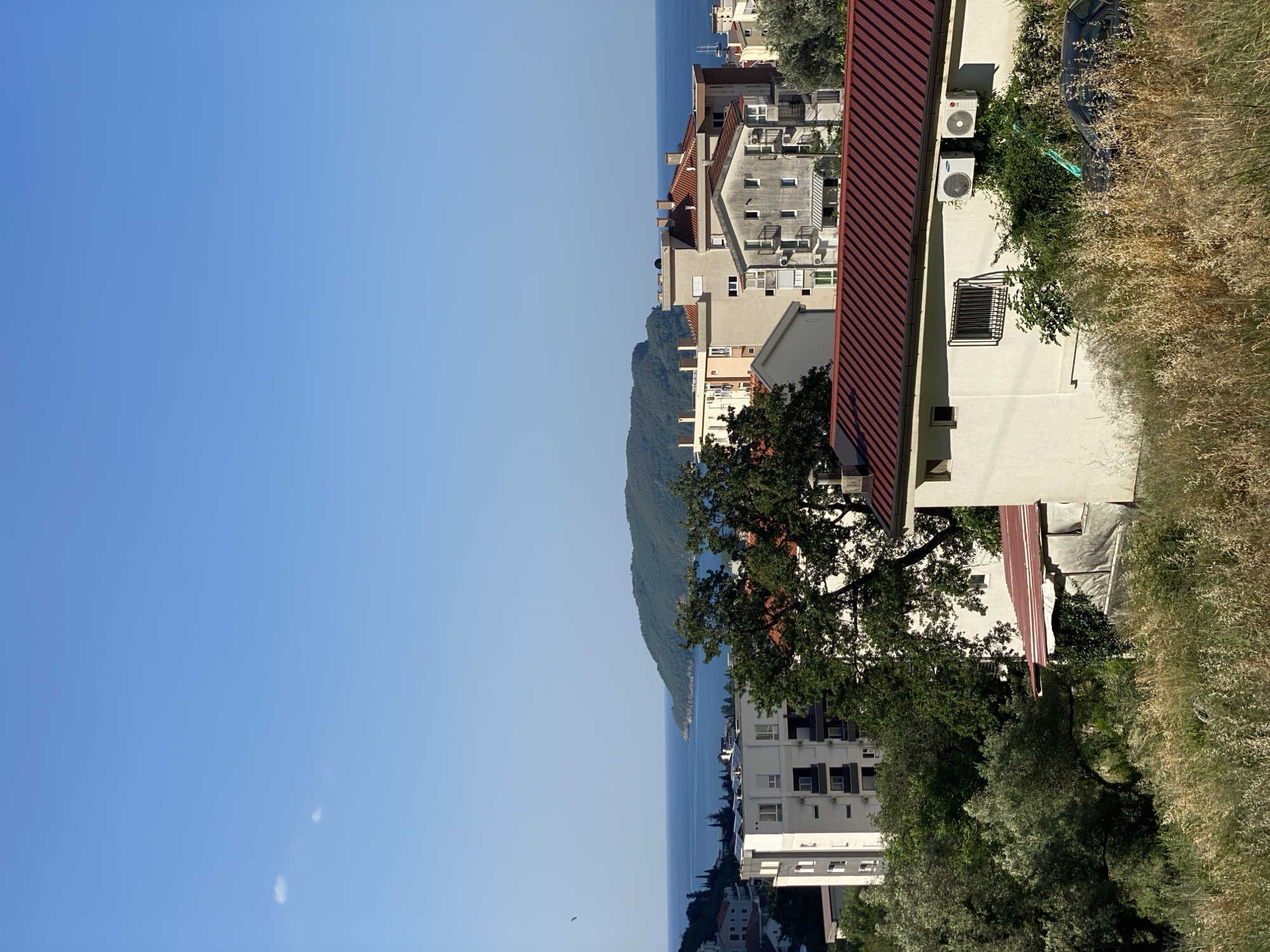 The image shows a panoramic view of a coastal city, likely Budva, Montenegro, with buildings of various architectural styles. In the foreground, there is a hillside covered with dry grass and lush green trees. Several residential buildings with white and beige facades are visible, some with red tiled roofs. In the middle ground, there are larger apartment buildings, some featuring balconies and windows. A body of water, likely the Adriatic Sea, stretches across the background, with a lush green island in the distance. The sky is clear blue with a few faint clouds. The scene appears to be captured during daylight hours, with bright sunlight casting shadows. There are no people or animals visible in the image. The environment suggests a Mediterranean coastal town, with dense vegetation and a clear view of the sea.