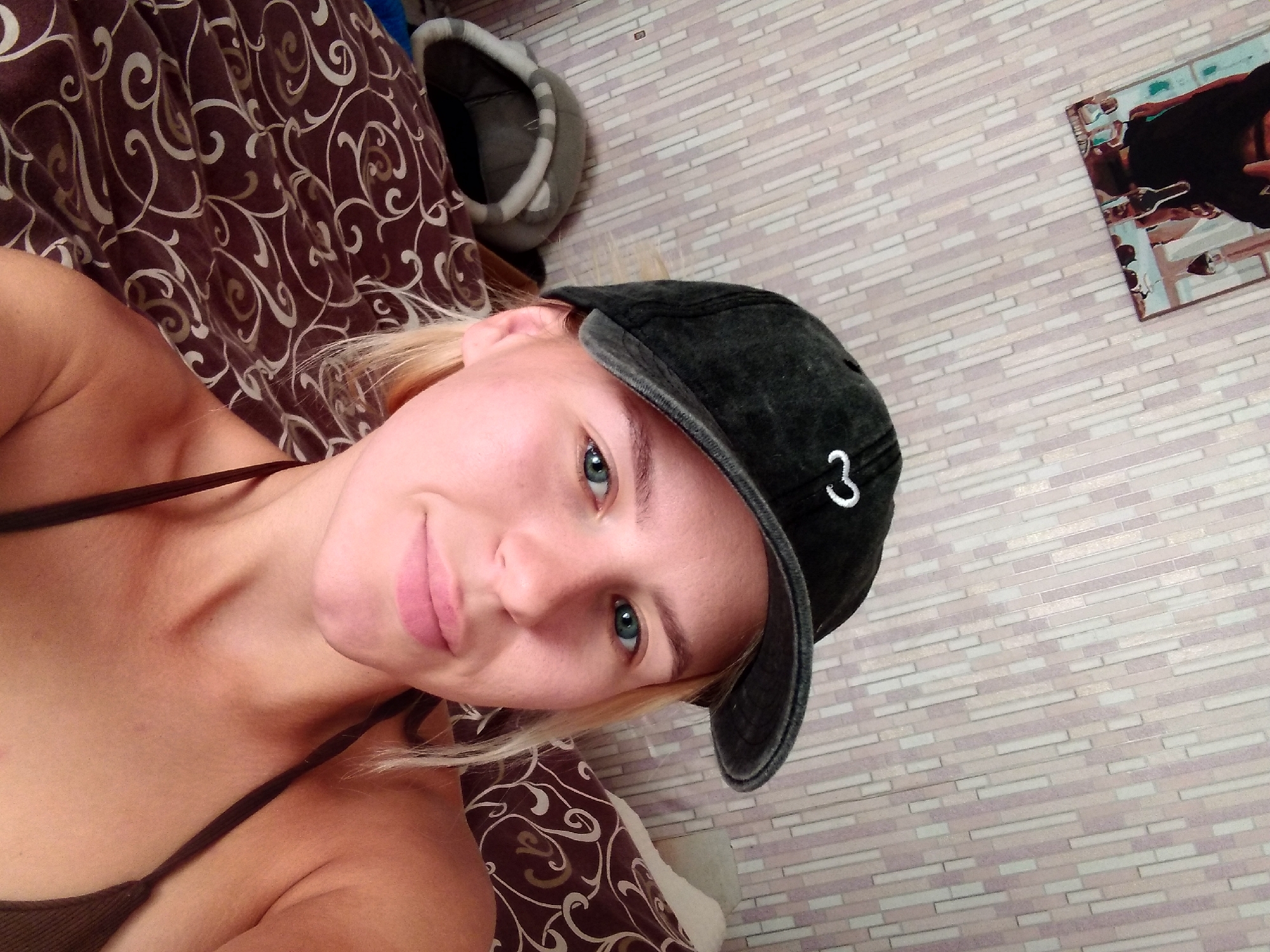 A young woman with blonde hair and blue eyes is looking at the camera. She is wearing a black baseball cap and a dark-colored bikini top. The woman is in a room with patterned wallpaper and a patterned blanket. There is also a cat bed in the background. The image was taken in Radisne, Ukraine.