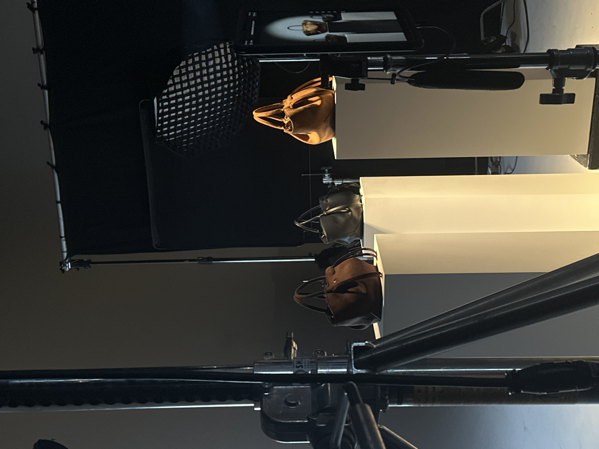 This image depicts an indoor product photography or videography setup in a professional studio, likely located in London, United Kingdom. The scene, captured with a vertical orientation, showcases three handbags arranged sequentially on white, rectangular platforms.
From top to bottom, the handbags include a light tan or camel-colored bag, a dark grey bag, and a dark brown or suede-textured bag, all appearing to be small-to-medium sized women's purses. An activity of product shooting is evident, with a monitor or tablet in the upper left displaying a product shot of the tan handbag, suggesting a live preview or recent capture.
Notable details include studio lighting equipment, with a large light modifier featuring a grid visible on the far left, indicative of controlled illumination. The background is predominantly dark, allowing the products and equipment to stand out. Various metal poles, stands, clamps, and rigging, typical of a studio environment, are present throughout the frame. A horizontal bar extends from the right, with a black oblong accessory (possibly a microphone or light bar) attached beneath it. A silver clamp near the bottom center shows partial text, possibly "SH". The overall impression is that of a commercial photoshoot for fashion accessories.
