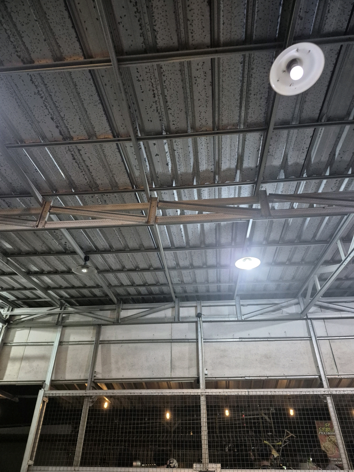 The image shows an interior view looking upwards at a metal roof structure in Pabelan, Indonesia. The roof is made of corrugated metal sheets supported by a framework of metal beams and struts. There are two industrial-style lights visible: one in the upper right quadrant and another in the middle right quadrant, both emitting a bright light. Below the roof structure, a wire mesh fence encloses an area where two bare incandescent bulbs are visible, casting a warm glow. In the background, through the fence and beyond the lit bulbs, the scene is dark, but there are hints of objects and possibly a poster or sign on the right side, including text that appears to be in Indonesian. The overall impression is of an industrial or utilitarian space, possibly an outdoor market or a covered workshop.