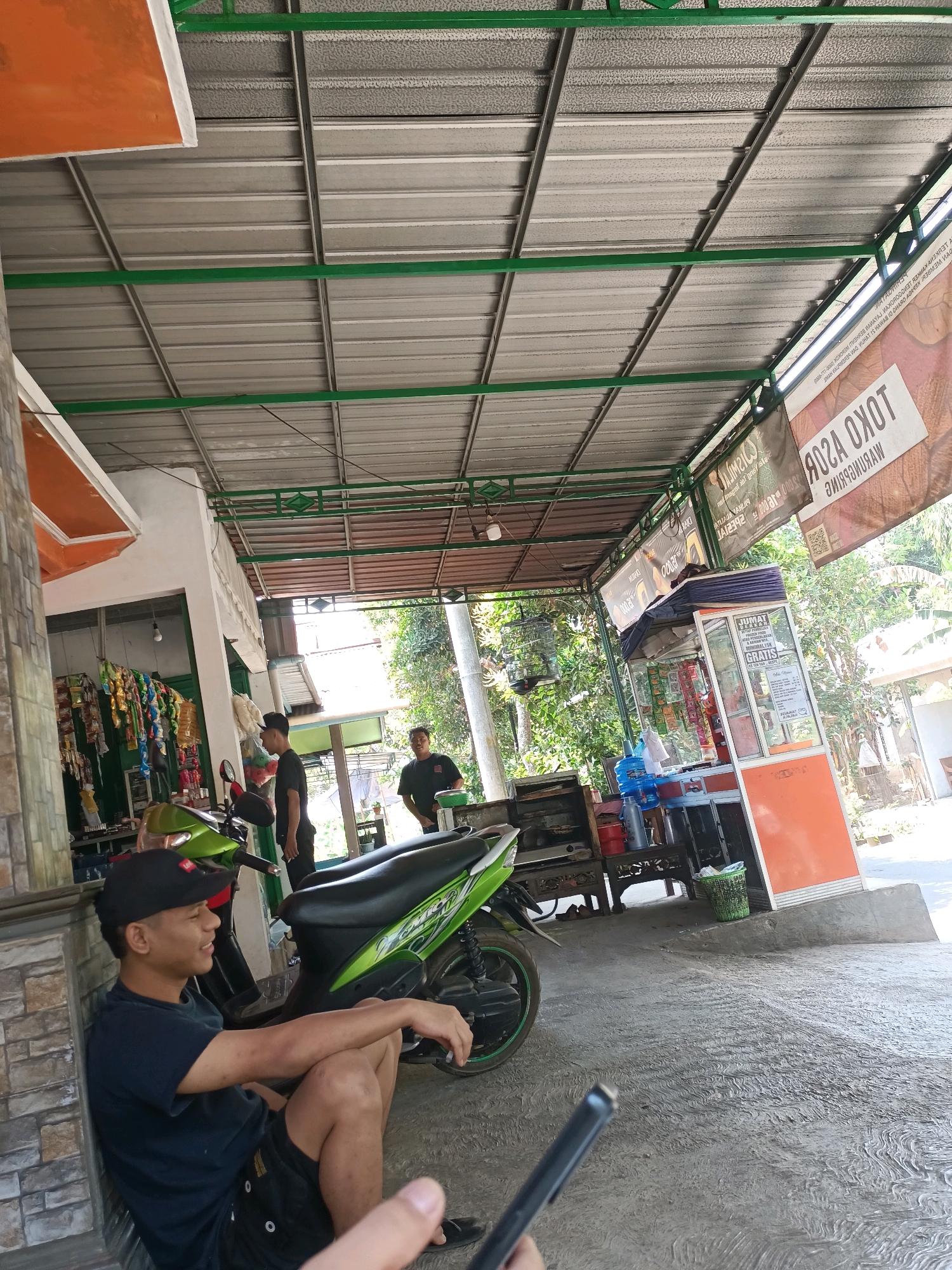 The image depicts a street scene in Warungpring, Indonesia, under a covered outdoor area. In the foreground, a young man wearing a black t-shirt, black shorts, and a black baseball cap is sitting on the ground, leaning against a brick wall. He is looking to his left and appears to be in a relaxed posture. A green and black motorcycle is parked next to him.
Behind the young man, two other men are standing. One man, dressed in a black t-shirt, stands near a small food stall with an orange and white facade. This stall displays various packaged goods and has a glass front. The other man, wearing a black t-shirt, is standing further back, near some trees and foliage.
The environment includes a corrugated metal roof supported by green metal beams, suggesting an open-air structure. Various banners are hung from the roof, including one with text that reads "TOKO ASOR WARUNGPRING." The ground is paved with concrete, and there are plants and trees visible in the background, indicating a somewhat rural or semi-urban setting. A birdcage is also visible hanging near the left side of the frame. The overall impression is a casual, everyday scene in a local business area.