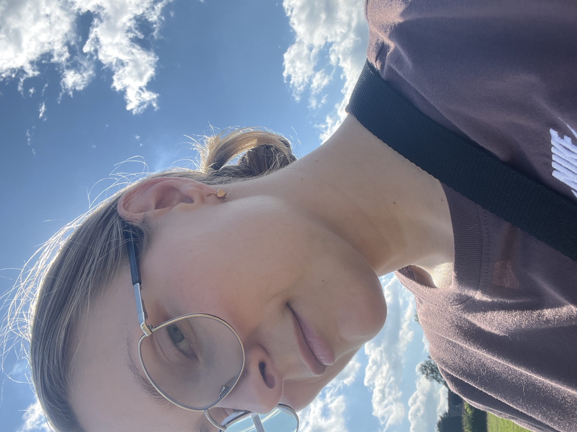 The image is a close-up, outdoor, selfie-style photograph of a person's face and upper torso. The subject is wearing large, round, gold-rimmed glasses and a simple, purple t-shirt with a white Nike logo visible on the chest. Their hair is pulled back in a low ponytail, and they are wearing a small, heart-shaped earring in their visible ear. The background consists of a bright blue sky with scattered white clouds, suggesting daytime and fair weather. There is a hint of green foliage and possibly some distant buildings in the lower left corner, implying an outdoor, possibly rural or park-like setting. The lighting suggests the sun is to the upper left of the frame, casting highlights on the subject's hair and skin. The image does not provide any specific visual cues to pinpoint the location within Saint Vith, Belgium.