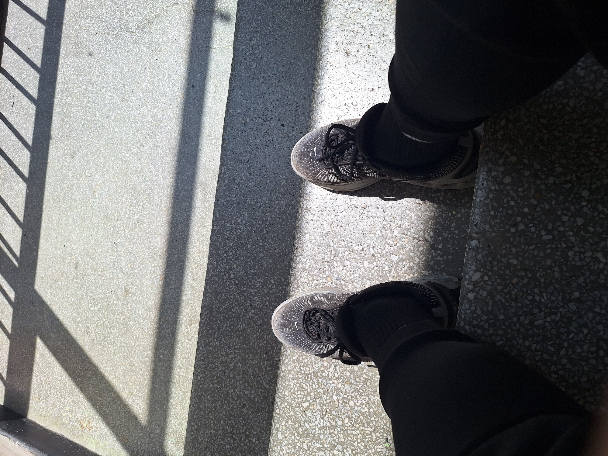 A first-person perspective shot shows a person's feet, clad in black socks and athletic shoes, resting on concrete steps. The steps have a speckled grey and white pattern. Sunlight casts diagonal shadows from an overhead railing onto the steps, creating a stark contrast between light and dark areas. The environment appears to be an outdoor public space, possibly a park or a stairway in an urban setting.