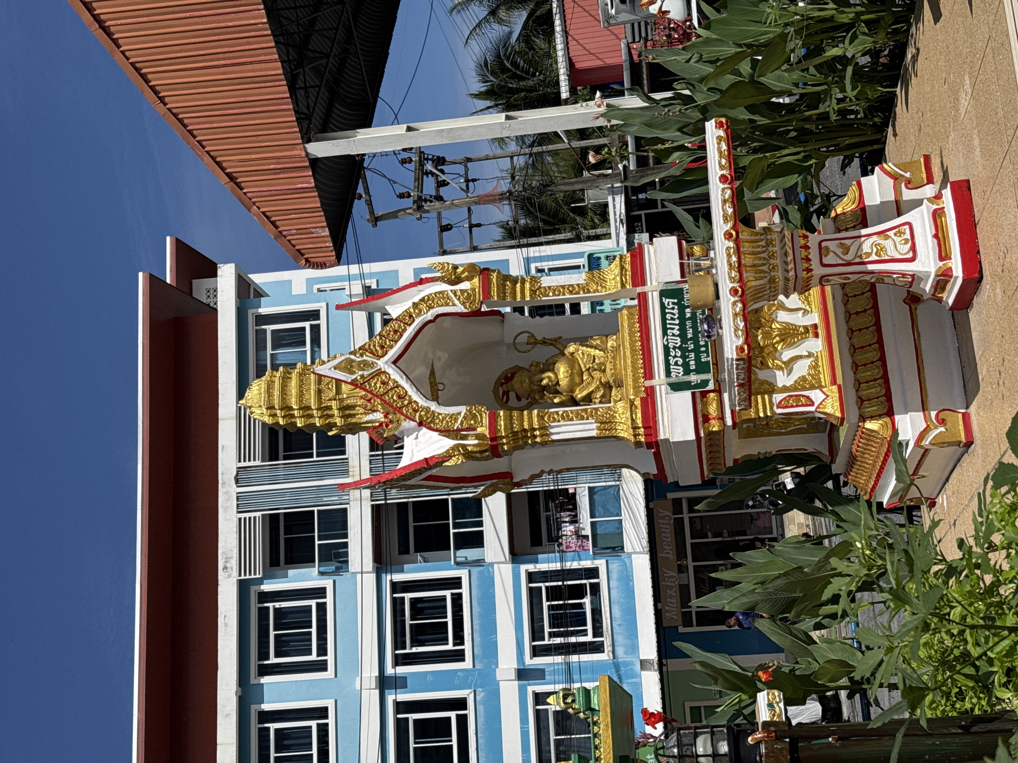 In Patong, Thailand, a small, ornate shrine, predominantly white and gold with red accents, stands in the foreground. Atop the shrine sits a golden statue of a deity, possibly Ganesh, with an elephant-like head. Below the deity, a green sign displays Thai script. The shrine rests on a patterned base and is surrounded by lush green foliage. Behind the shrine, a vibrant blue building with multiple windows and balconies, featuring a red roofline, dominates the background. Adjacent to the blue building is a structure with a corrugated red roof, supported by concrete pillars. Electrical poles and wires are visible, indicating an urban setting. Sunlight illuminates the scene, casting shadows and highlighting the bright colors. The overall impression is one of a tropical urban environment with a prominent religious or cultural element.