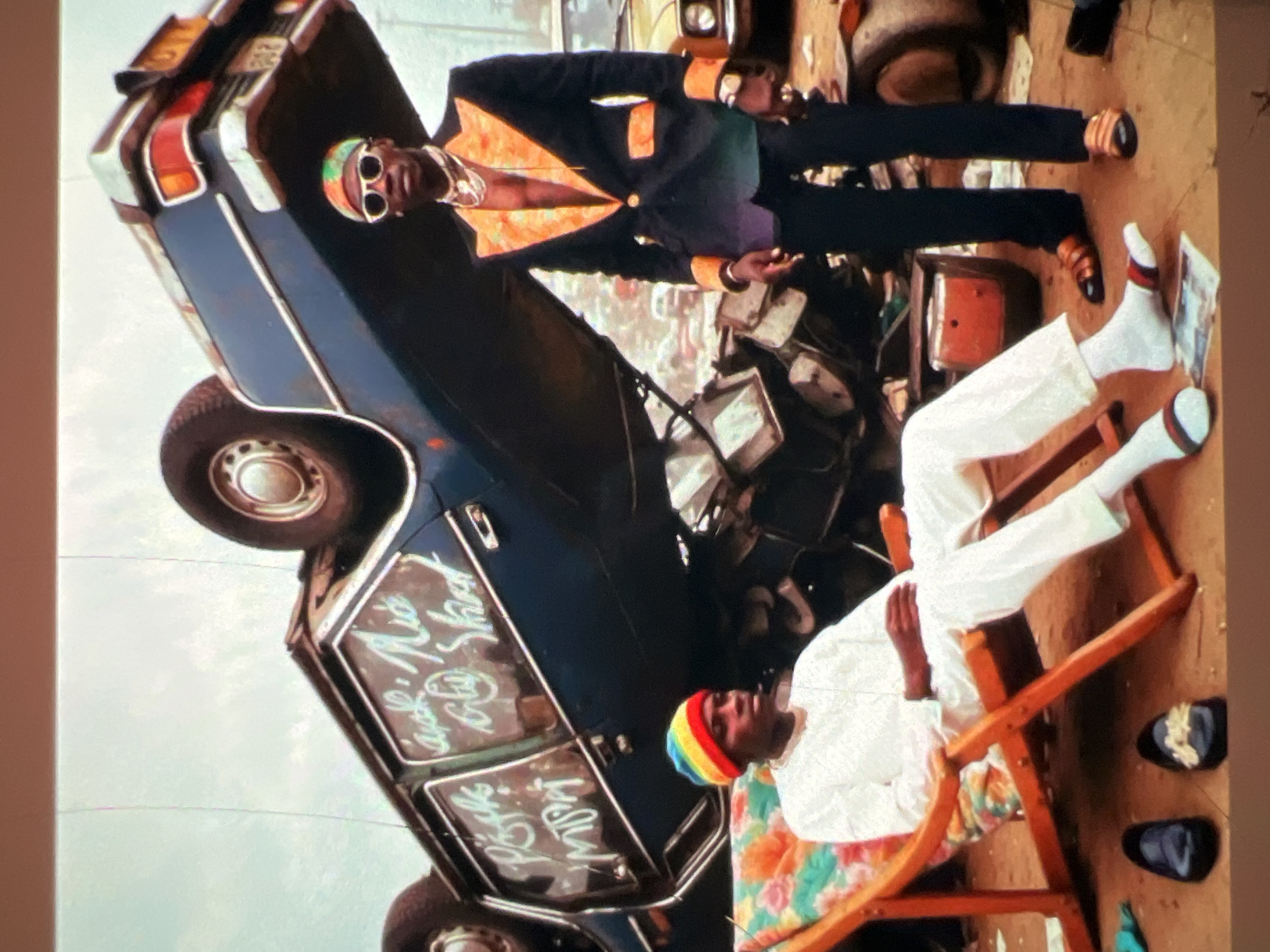 The image depicts a surreal outdoor scene, possibly in a junkyard or a less maintained area within Lagos, Nigeria. The most prominent object is a dark blue car, seemingly from the 1970s or 1980s, overturned on its side, with its wheels pointing upwards. The car's windows are scrawled with white graffiti.
In the foreground, two men are present. One man, seated in a wooden lounge chair adorned with a colorful floral cushion, is dressed in a white tunic and pants, and wears a rainbow-colored beanie. He appears relaxed, with his legs crossed. Next to him, on the ground, are a pair of dark loafers and another pair of ornate black shoes.
Standing behind the seated man is another man, dressed in a striking black suit with vibrant orange lapels and cuffs, open to reveal his chest. He wears sunglasses and multiple gold necklaces, and has a watch on his left wrist. He is looking towards the camera. His attire suggests a flamboyant style.
The environment is gritty and appears to be an open space with scattered debris and other vehicles in the background, some of which are partially visible and seem to be in a state of disrepair. The sky is overcast. The overall impression is one of an artistic and possibly staged scene, juxtaposing elements of decay with vibrant fashion.