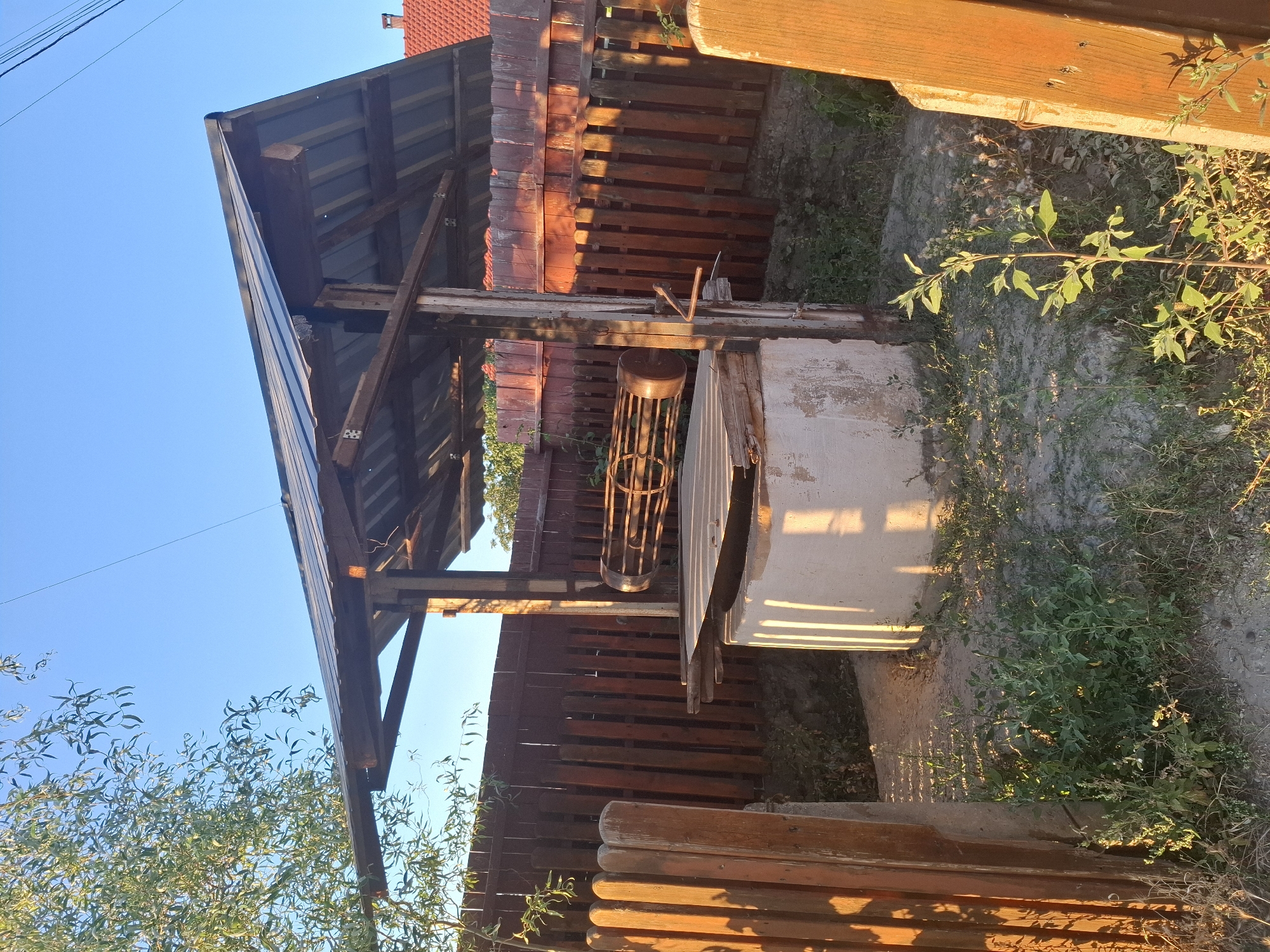 The image depicts an outdoor scene in Dubăsarii Vechi, Moldova, featuring an old, rustic well. The well itself is constructed of concrete and is partially covered by a wooden structure with a metal roof, suggesting it is a functional well with a protective shelter. A wooden winch mechanism, likely used for drawing water, is visible. The well is surrounded by a wooden fence, some parts of which appear weathered. Vegetation, including tall grass and small plants, grows around the base of the well and fence. The setting appears to be a backyard or a rural yard, given the presence of the fence and the unkempt nature of the vegetation. The lighting suggests it is late afternoon or early morning, with sunlight casting long shadows. There are no people or animals visible in the image.