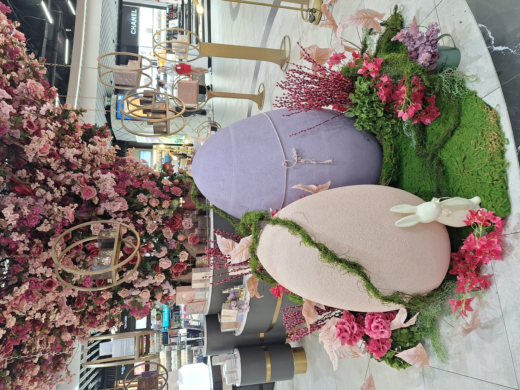 The image captures an elaborate Easter display inside a department store in Sofia, Bulgaria. Two oversized faux Easter eggs, one lavender and one pale pink, are arranged on the floor, decorated with moss, pink and purple flowers, and decorative branches. A small white ceramic bunny rests on the pale pink egg. The scene is heavily decorated with an abundance of pink and red flowers, creating a lush and festive atmosphere. In the background, shelves of perfume and cosmetics are visible, indicating the location is likely a beauty or fragrance section within the store. The store has black and gold fixtures. Metal chandeliers are installed above the cosmetic counters.