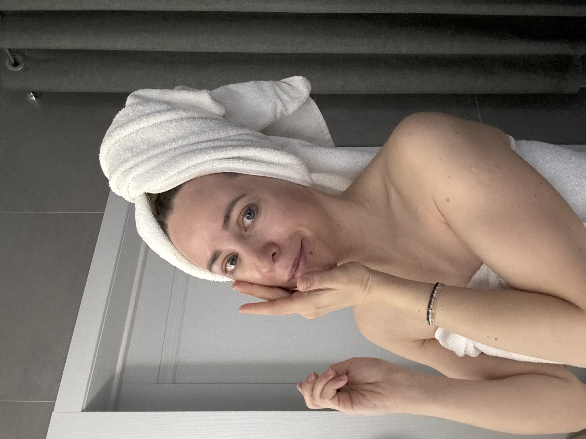 A person is lying down, wrapped in a white towel with their hair covered in a white towel. The person is looking at the camera with a slight smile. They are resting their cheek on their hand. The background consists of gray wall and a white wood-paneled structure. The scene is potentially set in the city of Chaikovsky, Russia.