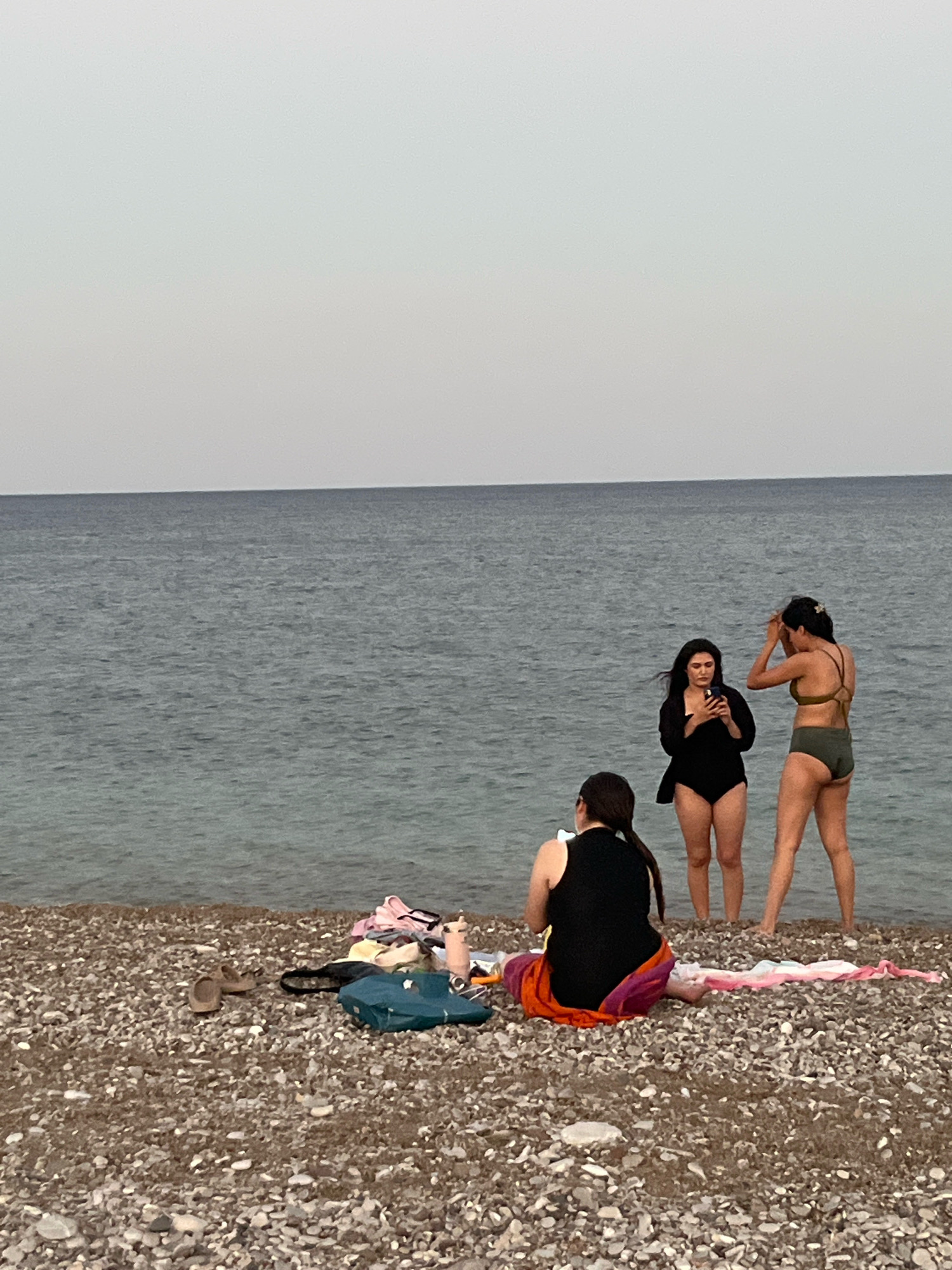 Three women are on a pebble beach with the sea in the background. The woman on the left is sitting on the beach, facing away from the camera, with a towel wrapped around her. The woman in the middle is standing and looking at her phone. The woman on the right is standing with her back to the camera, adjusting her hair. There are several items of clothing and a bag on the beach to the left of the sitting woman. The scene is in Kemer, Turkey.