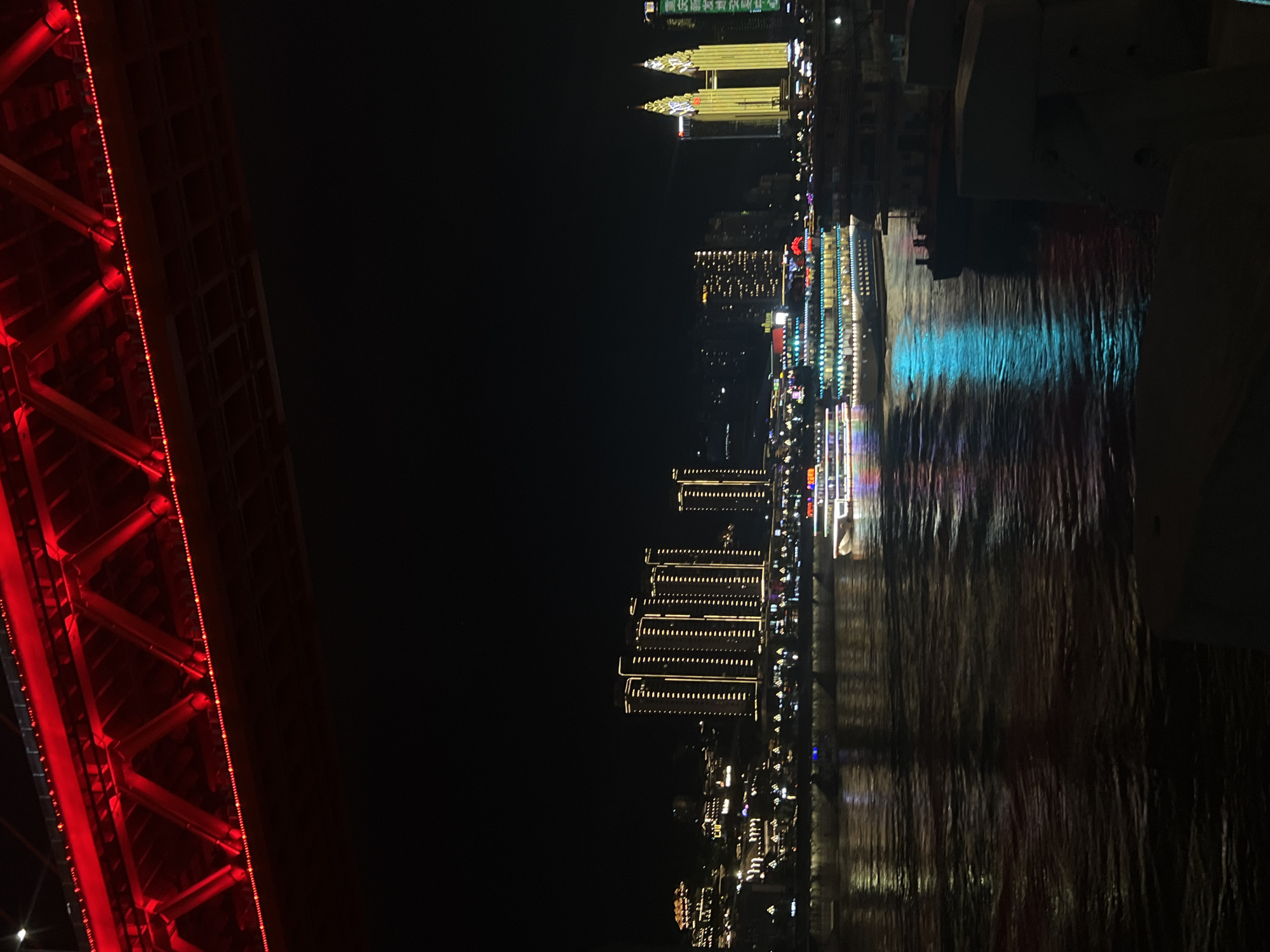 The image captures a night scene on a river in Yuzhong District, China, likely the Yangtze River. A brightly lit suspension bridge, adorned with red lights, spans across the top of the frame. Below the bridge, a wide river flows, reflecting the vibrant lights of the cityscape and passing boats.
On the river, a multi-decked passenger boat is illuminated with colorful lights, cruising gently on the water. Its reflection shimmers on the dark surface of the river, creating streaks of cyan and magenta. In the background, the Yuzhong District skyline is visible, characterized by numerous tall buildings illuminated with warm, yellow lights. Two distinctive skyscrapers with pointed, illuminated tops stand out prominently. The buildings suggest a bustling urban environment. The overall atmosphere is one of vibrant nightlife and urban illumination against the dark canvas of the night sky.