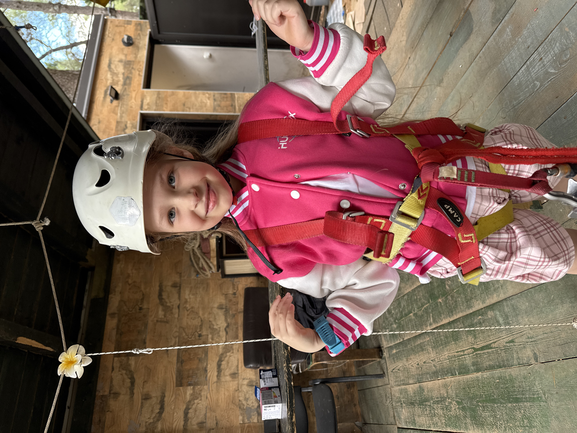 A young girl is wearing a white helmet and a red harness with the word "CAMP" on it, over a pink jacket with white stripes, while standing on a wooden structure. A white flower is tied to a rope nearby. Based on the style of construction, the location may be an adventure park or recreational climbing area. While the style of wooden construction is consistent with many outdoor recreational facilities, there is not sufficient information to specifically identify the location in Podgorica, Montenegro.