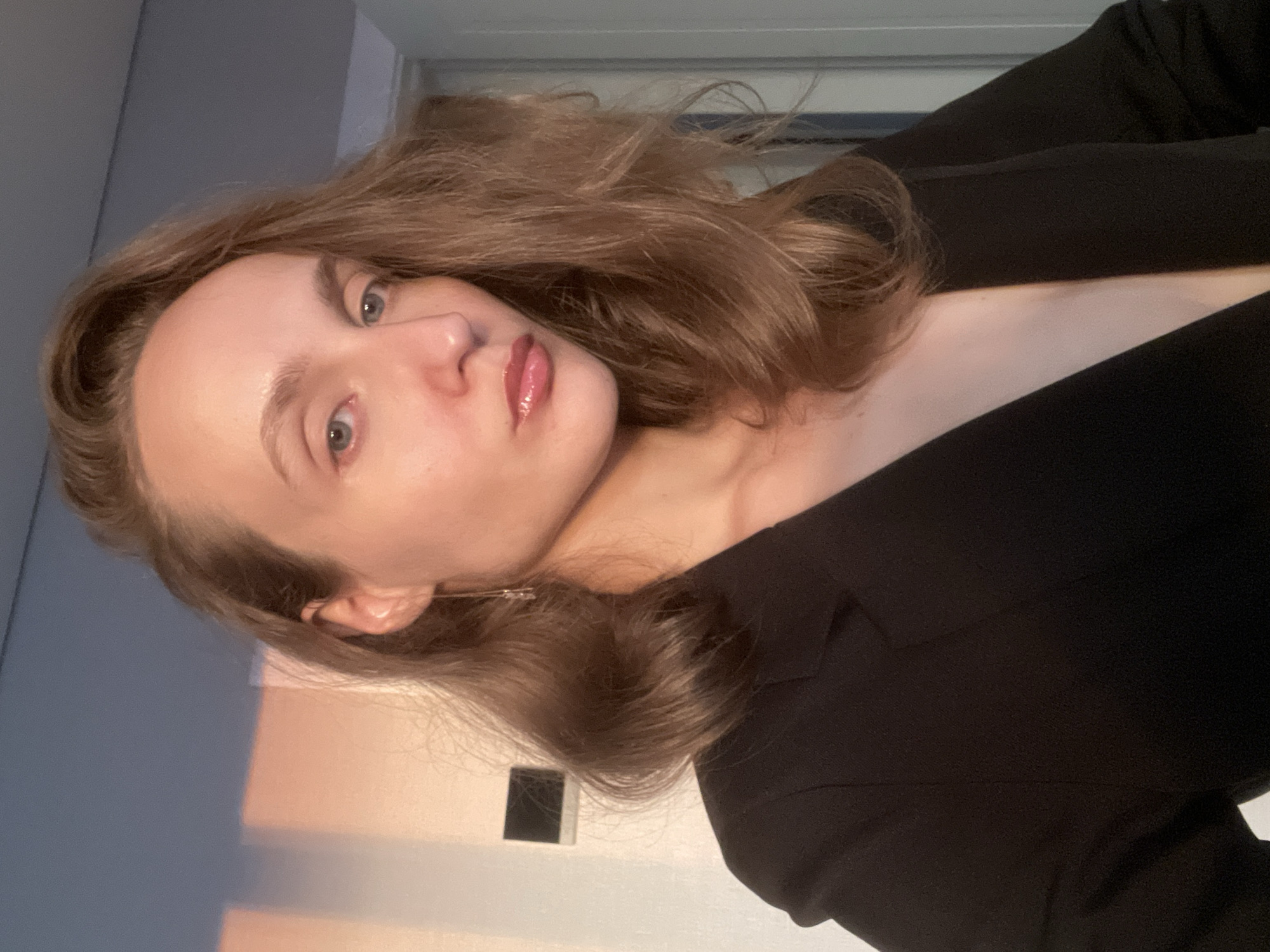 A medium shot captured from a slightly low angle shows a young woman with light brown hair styled loosely around her shoulders. She is wearing a black blazer that is open, revealing her décolletage. The woman has light blue eyes, well-defined eyebrows, and glossy pink lips. She is looking slightly away from the camera, with her head tilted. Her left ear is visible and adorned with a long, delicate earring.
The background consists of a wall that transitions from a light blue at the top to a warm, peachy-orange hue lower down, suggesting the play of light. A dark, rectangular object, possibly a light switch or an outlet cover, is mounted on the wall to the left of the woman. To the right, a door frame and part of a door are visible. The overall lighting suggests it might be late afternoon or early evening, with a soft, warm light casting a glow on the wall and the woman's face.
The image does not contain any specific visual cues to identify the location as Podshivalovo, Russia.