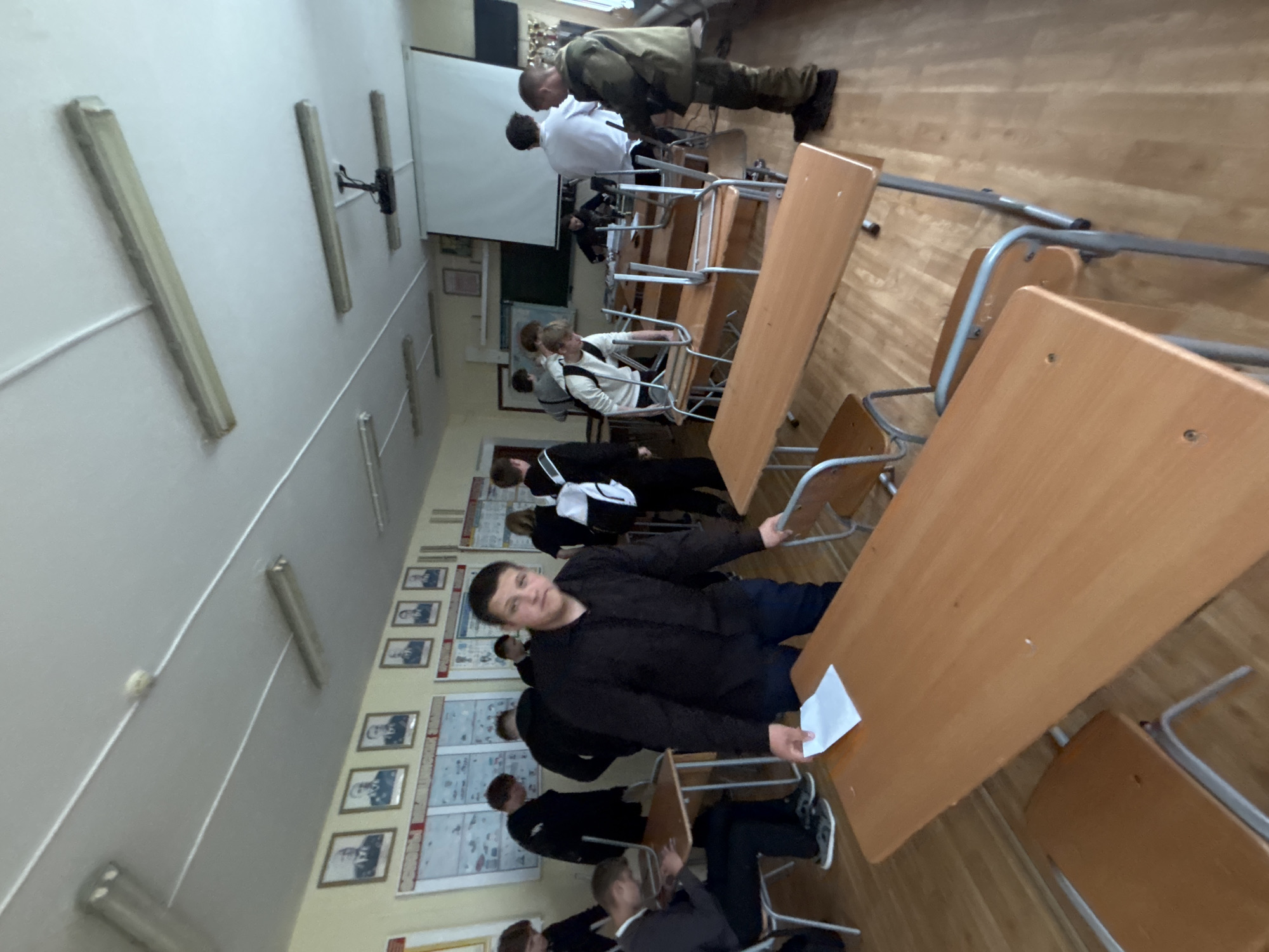 The image, rotated vertically, depicts an indoor classroom or lecture hall. Several young men or boys are present, some seated at wooden desks with metal chairs, others standing.
In the background, near the front of the room, a man in a camouflage military-style uniform, including a jacket, pants, and boots, stands beside a desk. Another young man in a white shirt is also standing in that area, facing away from the camera. A large white projector screen is visible on the front wall.
In the foreground and midground, students are seated at desks. One young man in a black long-sleeved shirt and dark pants is prominently featured in the midground, looking towards the camera with a neutral expression. He is holding a piece of white paper in his left hand. Other students are visible, some wearing white shirts, black shirts, or grey hoodies.
The room features a wooden floor. The walls are light-colored and have several fluorescent light fixtures mounted on the ceiling. Along one wall, framed portraits, possibly of individuals or historical figures, are hung interspersed with educational charts or posters, though no text is legible. A projector is mounted on the ceiling near the front. The overall atmosphere suggests an educational setting, possibly a class involving military or historical themes given the uniform and wall decor.