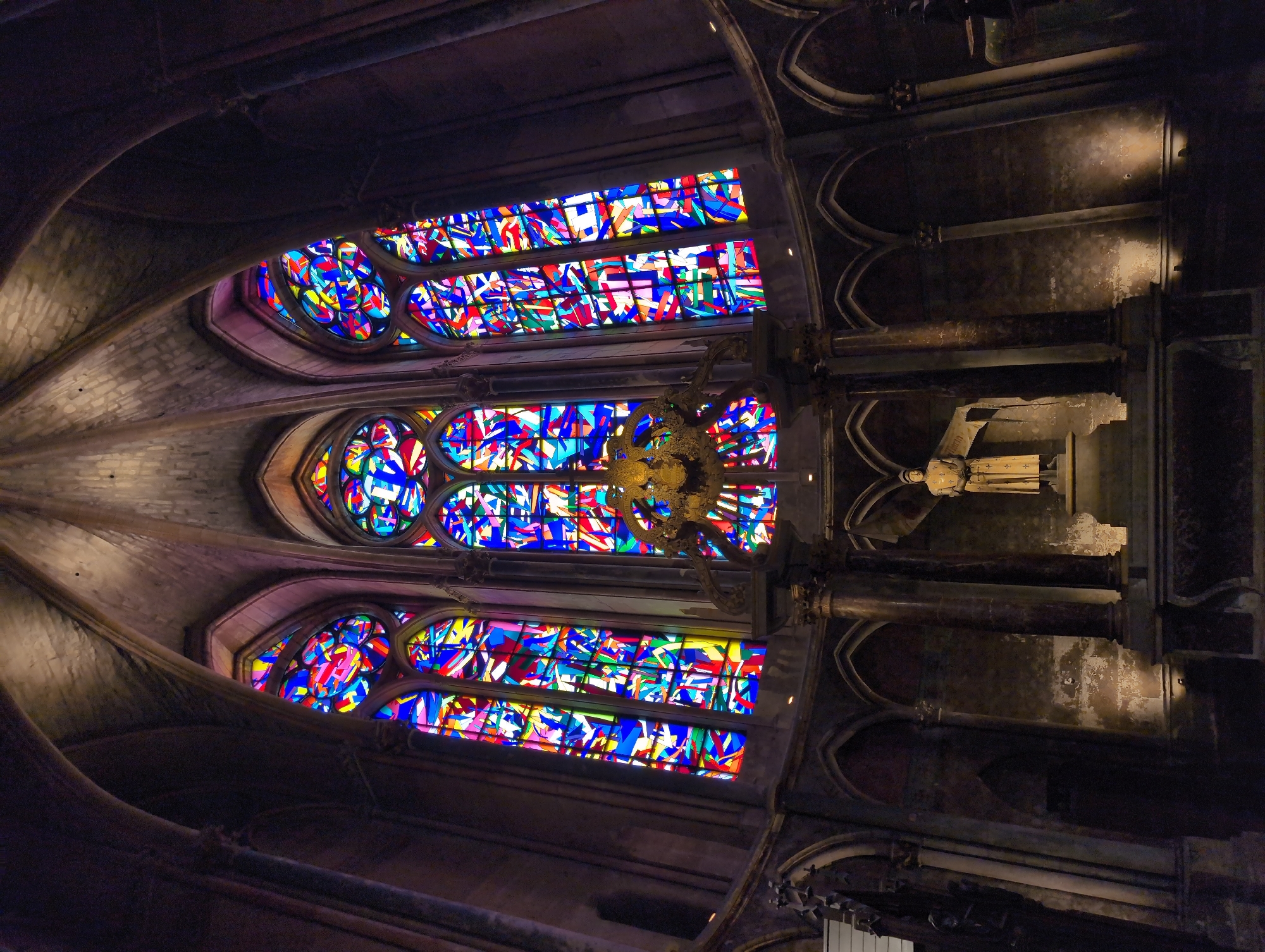 This is a wide-angle, low-light, interior shot of a cathedral, specifically the church of Saint-Germain in Urvillers, France. The focal point is a large, ornate stained-glass window filling the upper portion of the frame. The window is composed of numerous small, irregularly shaped panes of glass in vibrant colors, including red, blue, yellow, and green, set against a darker background. Below the window, a statue of a robed figure, possibly a saint or historical person, stands on a pedestal. The statue is illuminated by spotlights from the floor, casting shadows. The surrounding architecture features dark stone columns, arches, and vaulted ceilings, characteristic of Gothic architecture. The overall impression is one of grandeur and historical significance, with the colorful stained glass providing a stark contrast to the dark stone.