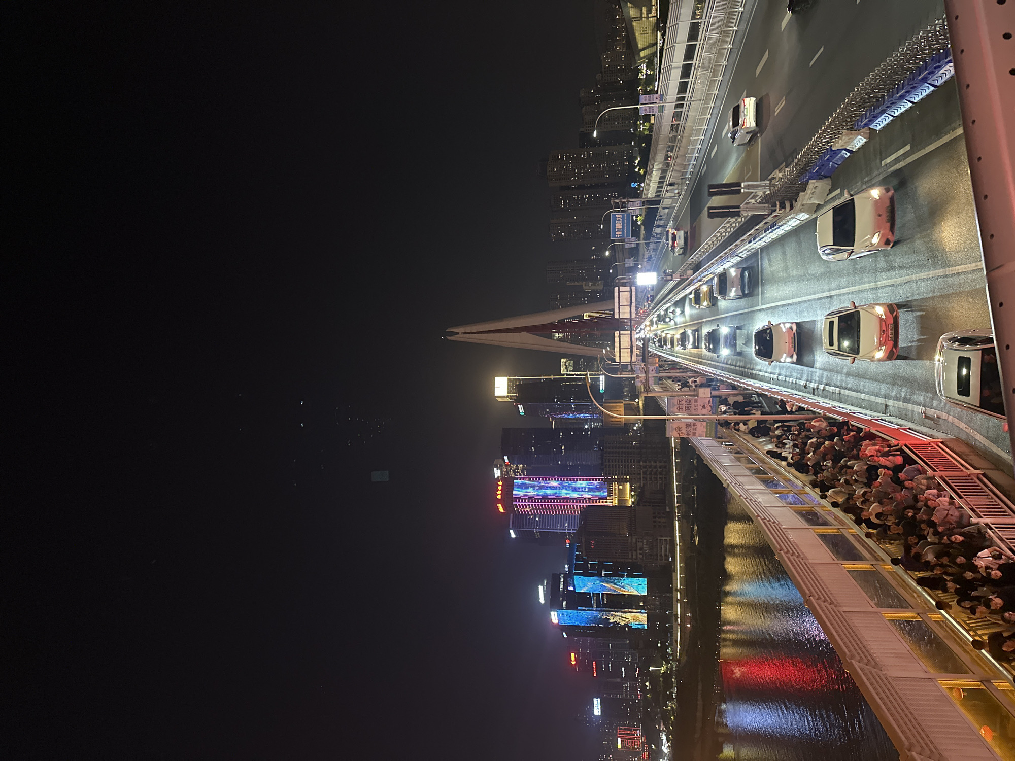 The image displays a lively nighttime urban scene, likely captured from an elevated position on a bridge in Yuzhong District, China. The primary subjects include a densely packed pedestrian walkway, vehicular traffic, and an illuminated cityscape.
A wide bridge spans a body of water, likely the Jialing River, with numerous cars visible on its roadway, their headlights and taillights glowing. A large crowd of people fills the pedestrian path on the bridge, suggesting a popular viewing spot or a public event. Their collective presence creates a bustling atmosphere, with individuals standing and walking, observing the surroundings.
In the background, a sprawling cityscape unfolds with numerous high-rise buildings and skyscrapers brightly lit, some featuring colorful decorative lights and large digital displays. A distinct cable-stayed bridge pylon is visible in the distance. The river below reflects the vibrant city lights, showing shimmering patterns of red and blue.
The scene is set at night under clear skies, creating a dynamic and energetic ambiance. Visible Chinese text includes "全阅," "民壶," "帐篷 阅读," and "读节 活动周," potentially indicating a "Reading Festival Activity Week" is taking place. The most significant textual clue is "千局门嘉陵江大桥," which is an OCR rendition of "Qiansimen Jialing River Bridge," a prominent landmark in Chongqing's Yuzhong District, explicitly confirming the location.