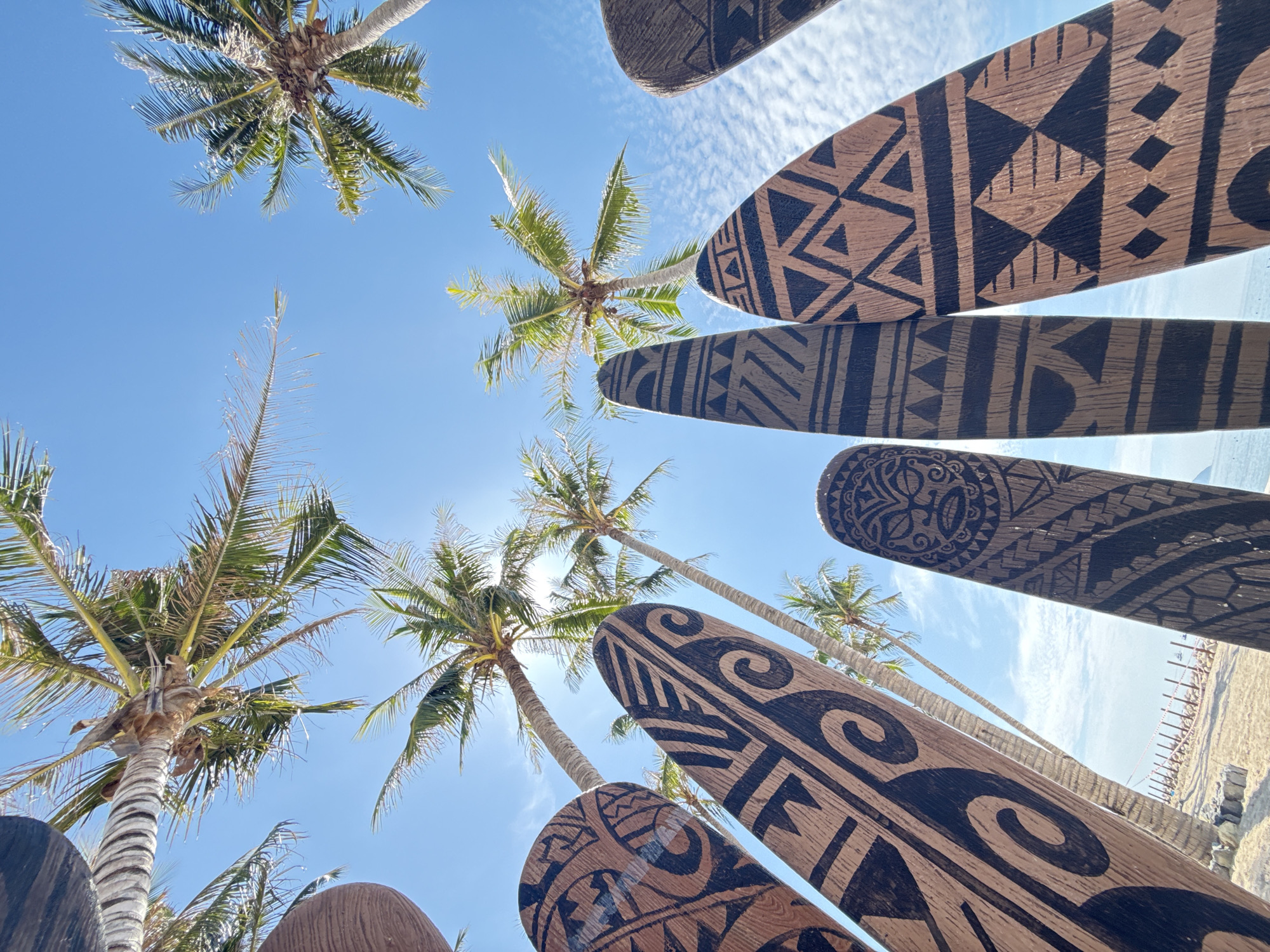 The image shows a low-angle, outdoor view of several carved wooden boards with Polynesian-inspired designs, standing upright on a sandy beach. Palm trees with green fronds are visible against a bright blue sky with scattered white clouds. The sun is shining, casting a lens flare on the right side of the image. The boards are spaced apart, with some of them angled towards the viewer. The sand is light brown, and a thin strip of water can be seen in the background, with a faint line of poles and some distant structures. This scene is likely on a beach in Nha Trang, Vietnam, given the presence of palm trees and the tropical atmosphere. There are no people or animals visible in the frame.