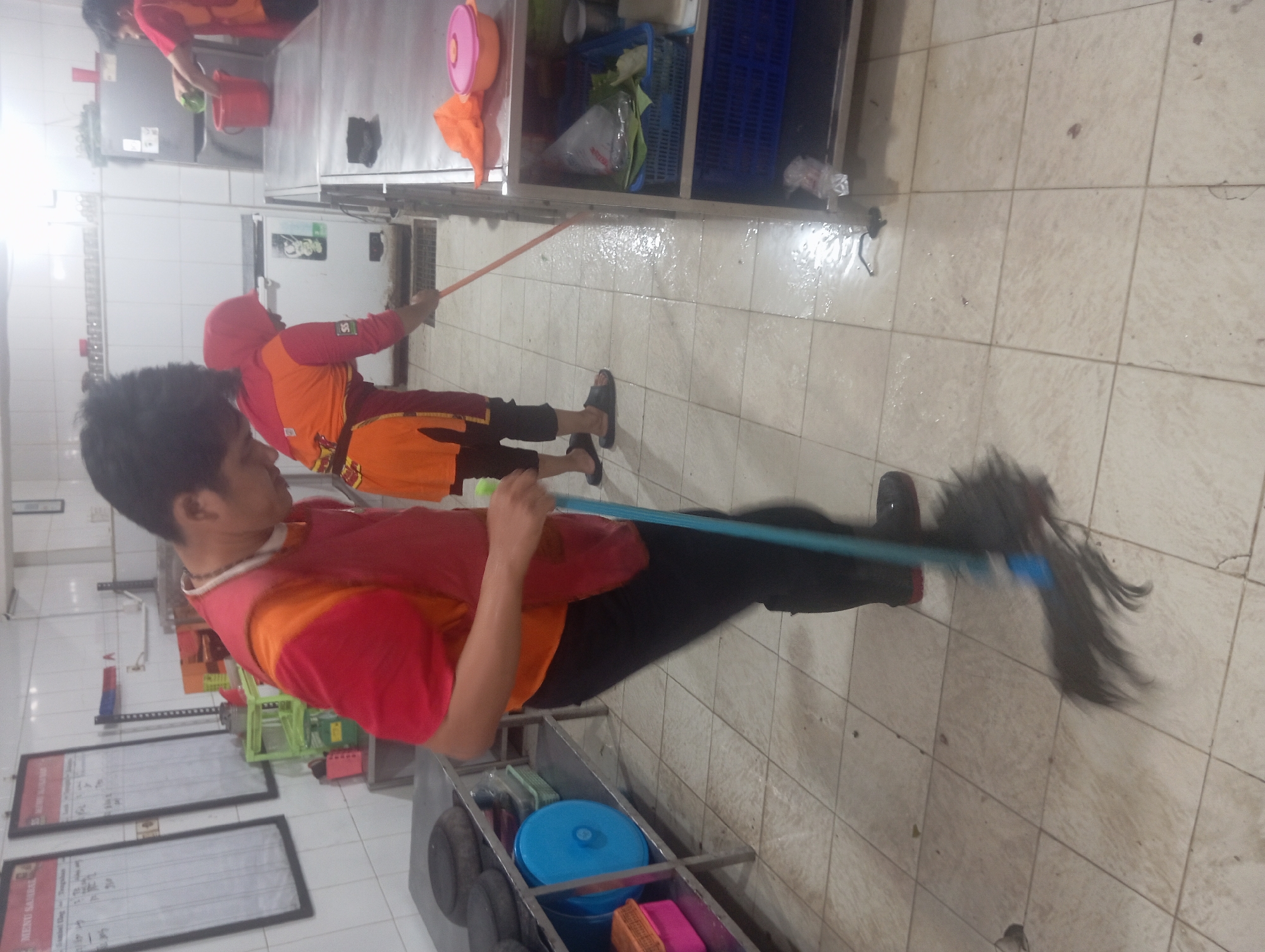 Two individuals are cleaning a kitchen or food preparation area. The person in the foreground, wearing a red shirt and an orange apron, is mopping the tiled floor with a dark mop. To the left, a metal shelf unit holds various items including round black objects, a blue container, and pink containers. In the background, another person, wearing a red shirt and an orange hooded garment, is also mopping the floor with an orange mop. This individual is near a stainless steel counter and a red bucket. The environment appears to be an indoor commercial kitchen, with white tiled walls and lighting fixtures visible on the ceiling. The overall scene depicts a cleaning operation in progress.