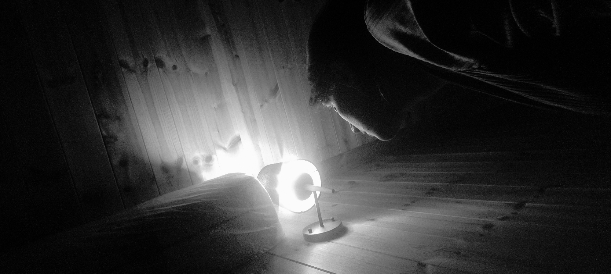 A monochrome photograph shows a dimly lit room with a wooden wall on the left and a wooden floor. A person is hunched over, their face partially obscured in shadow, peering downwards. A small, illuminated desk lamp sits on the floor, casting a bright light that washes out the details around it. The room appears to be indoors. There is no evidence to support the image was taken in Dunnae-myeon, South Korea.