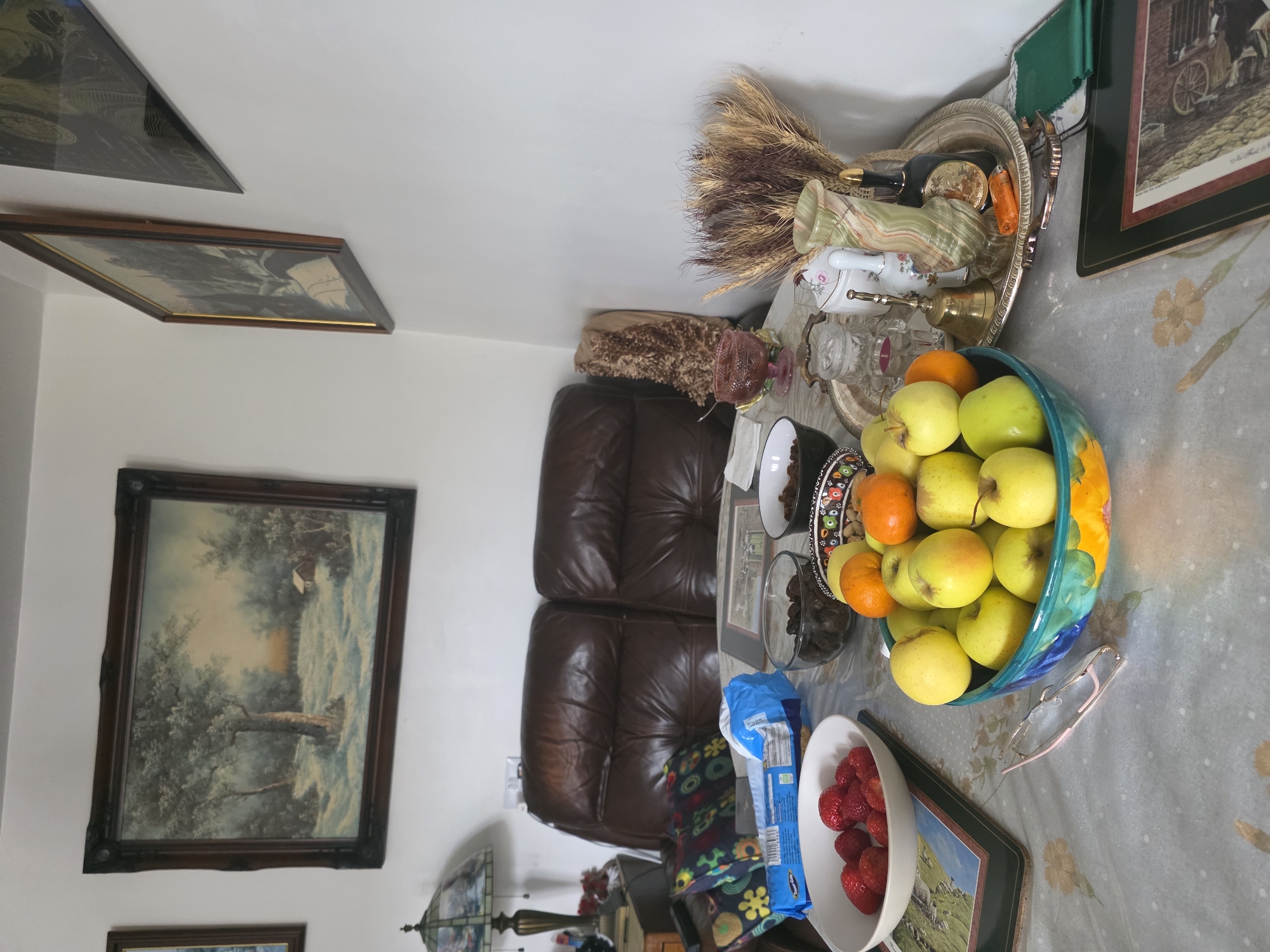 This interior image, set in Cwmbrân, United Kingdom, depicts a warm and decorated living room or sitting area. The scene is dominated by a table covered with a light-colored tablecloth, possibly grey with subtle floral patterns, which hosts a variety of items.
In the foreground, a large, vibrant teal-and-yellow bowl is filled with numerous green and yellow apples and a couple of oranges. To its left, a white bowl holds fresh red strawberries, and next to it, a blue plastic bag of what appears to be packaged food is visible, though the text on it is not clearly legible. A pair of light-framed eyeglasses rests on the tablecloth near the main fruit bowl.
Further back on the table, a clear glass bowl contains dried dark fruits, possibly dates or prunes. A decorative silver tray is arranged with several items, including a tall green variegated marble-like vase, a smaller white vase with a floral pattern, a brass bell, a clear glass condiment holder, and an orange lighter. A bundle of dried wheat stalks protrudes from behind these items.
The background features a comfortable-looking brown leather sofa or armchair with two visible cushions. The walls are adorned with framed artwork. On the left wall, a large landscape painting depicts trees and a small structure in a serene, perhaps wintry, setting. Above it, another smaller framed picture is partially visible. On the right side of the image, another framed artwork shows a rural scene with what appears to be a horse-drawn cart. A decorative lamp with a stained-glass shade is also partially visible in the lower-left corner, near the sofa. The room appears well-lit, suggesting either natural daylight or ample artificial lighting. No people are present in the image.