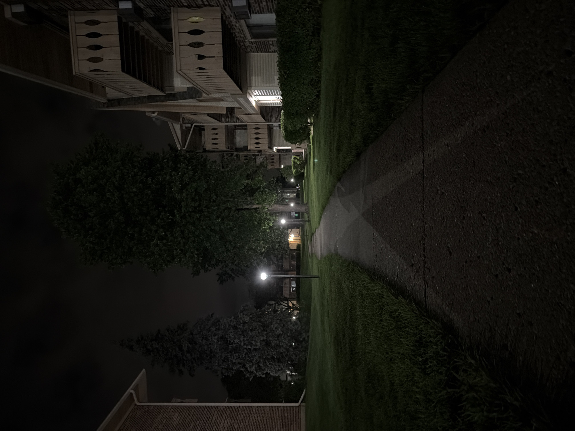 A paved walkway curves through a grassy area at night, illuminated by streetlights. To the right, a multi-story brick apartment building with balconies is visible. To the left, another brick building and several trees are present. The sky is dark and overcast. The location is the city of Vernon Hills, United States of America.
