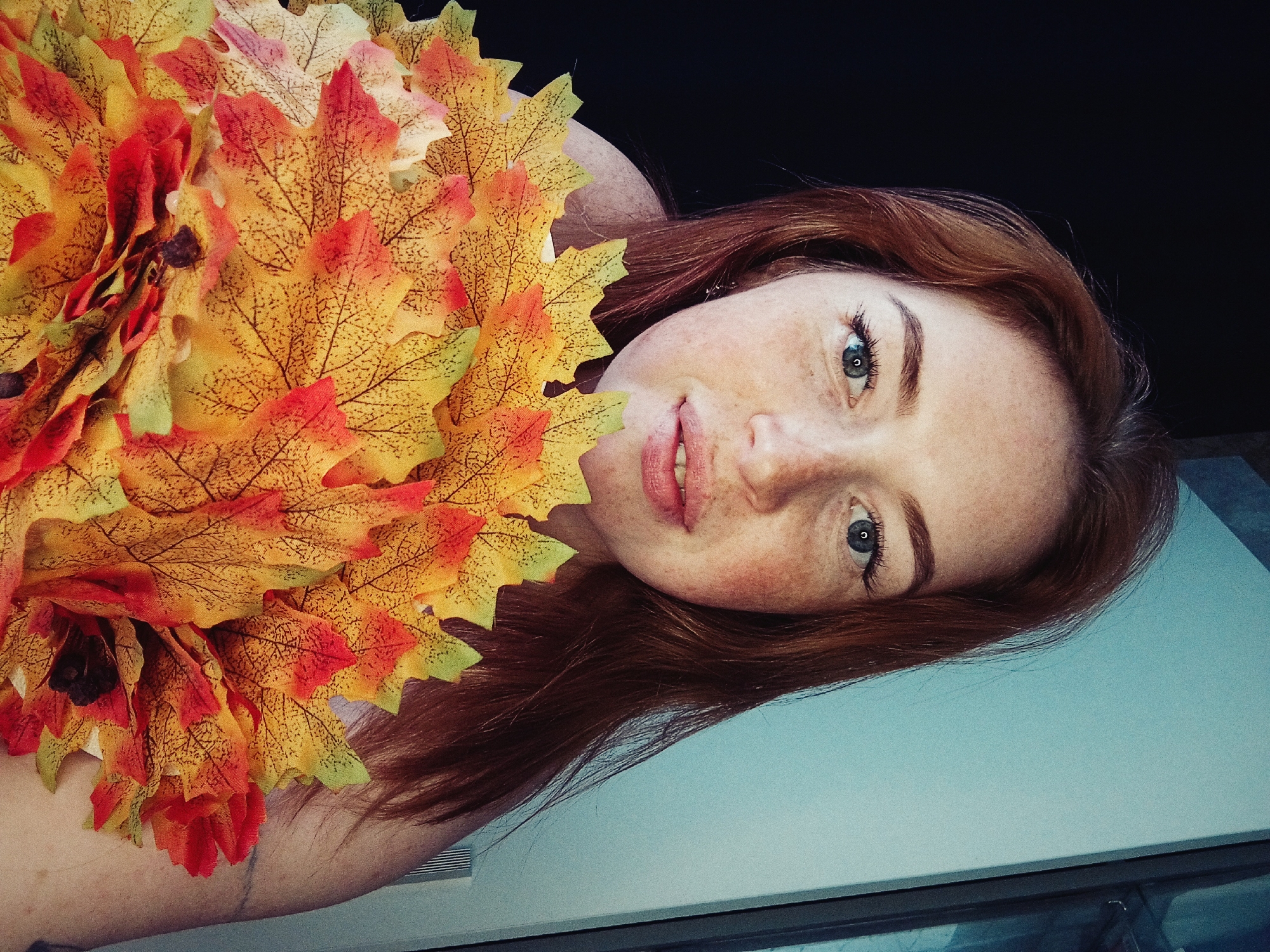 A fair-skinned, red-haired woman with freckles lies on her side. The location is unknown. She is surrounded by artificial autumn leaves in shades of yellow, orange, and red. The background is dark, possibly black. The surface she lies on is light blue and reflects some light. Her eyes are blue-green. Her lips are slightly parted.