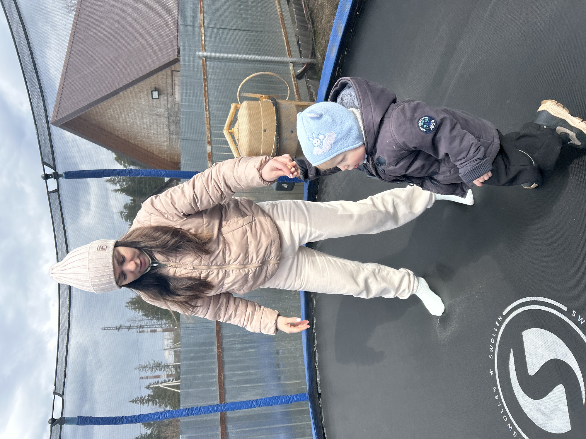 The image captures a moment between a young woman and a small child on an outdoor trampoline. The woman, with long brown hair, is dressed in a light beige puffer jacket, cream-colored pants, and a matching knitted beanie. She stands on the black trampoline mat, holding the hand of the child. The child is seen leaning forward on the mat, wearing a dark blue puffer jacket with a patch displaying "NAVY", dark trousers, and a light blue beanie adorned with a white embroidered bunny design. The trampoline features a blue padded edge and a safety net. In the background, under an overcast sky, elements of an outdoor environment are visible: a building with a brown roof, a yellow concrete mixer, a corrugated metal fence, and distant trees. The trampoline mat has a white logo and the word "SWOLLEN" printed on it. The scene suggests a supportive and playful interaction between the two individuals in what appears to be a cool, daytime setting, consistent with an outdoor play area in Chaikovsky, Russia.