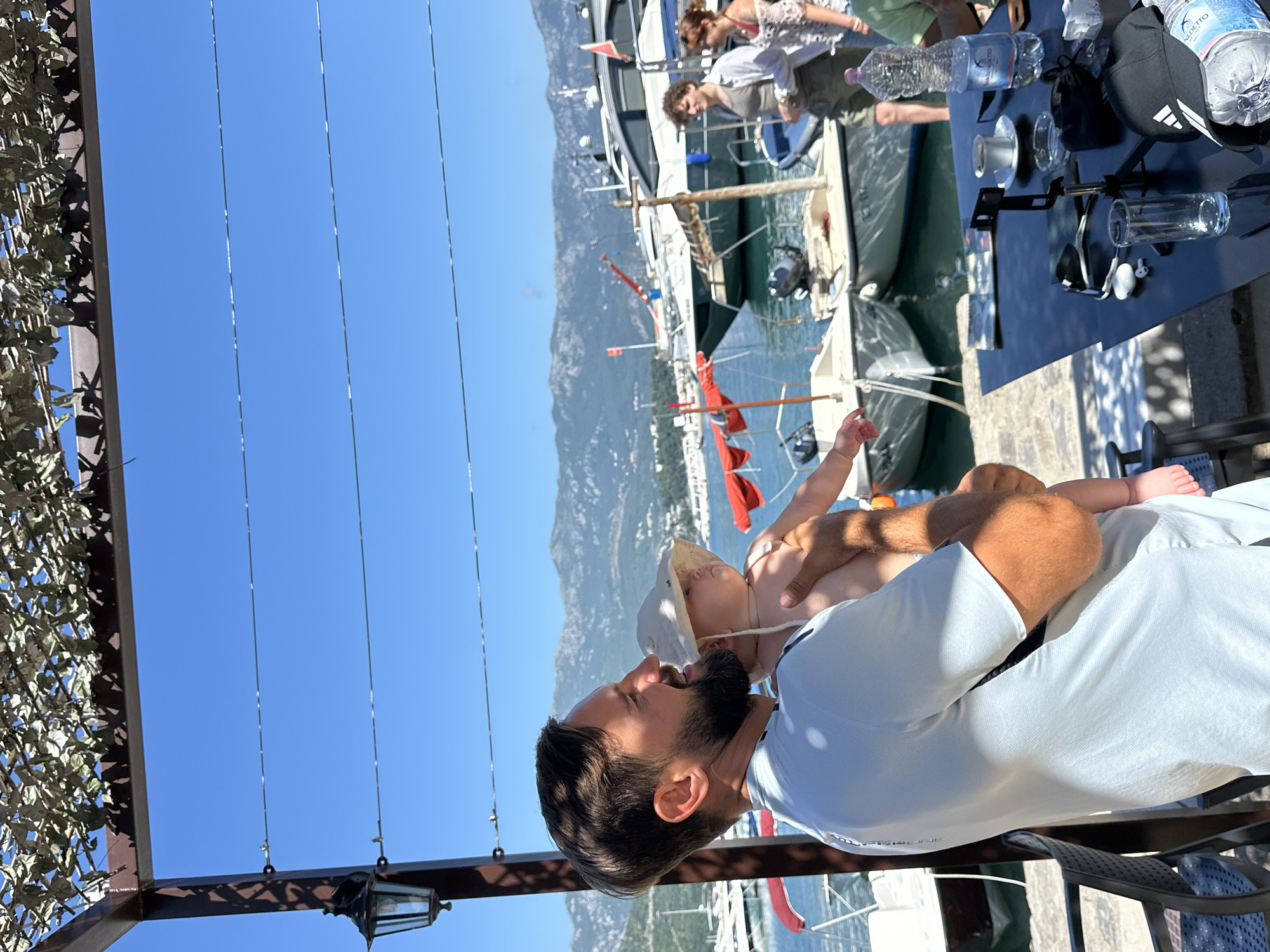 A man, likely in his 30s or 40s, with dark hair and a beard, holds a baby wearing a sun hat. They are positioned outdoors at what appears to be a waterfront restaurant or cafe with a view of boats docked in the harbor and mountains in the distance. The man is wearing a light-colored t-shirt, and the baby is partially visible, wearing only a diaper.
In the background, several boats, including a large yacht, are moored in the water. Other people are visible, including a woman in a white top and shorts, and a man in a light shirt and shorts. A black metal table in the foreground is set with drinks, a coffee cup, an ashtray, a cell phone holder, and a baseball cap. The scene is illuminated by bright sunlight, suggesting a warm, clear day. The presence of the boats, the harbor, and the mountainous backdrop strongly suggest the location is Budva, Montenegro, a coastal city known for its picturesque harbor and surrounding natural beauty.