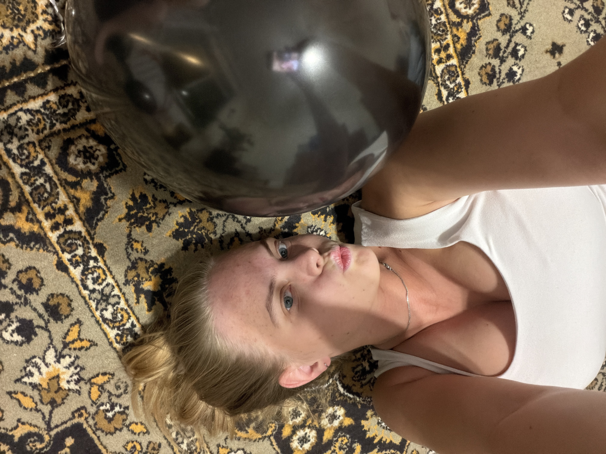 A woman with blonde hair and blue eyes is lying on a patterned rug. She is wearing a white tank top and a silver necklace. A black balloon is positioned above her face. The rug is predominantly beige, brown, and black. The photograph appears to have been taken from a high angle, looking down at the woman. The location in the city of Chaikovsky, Russia, cannot be determined from the image.