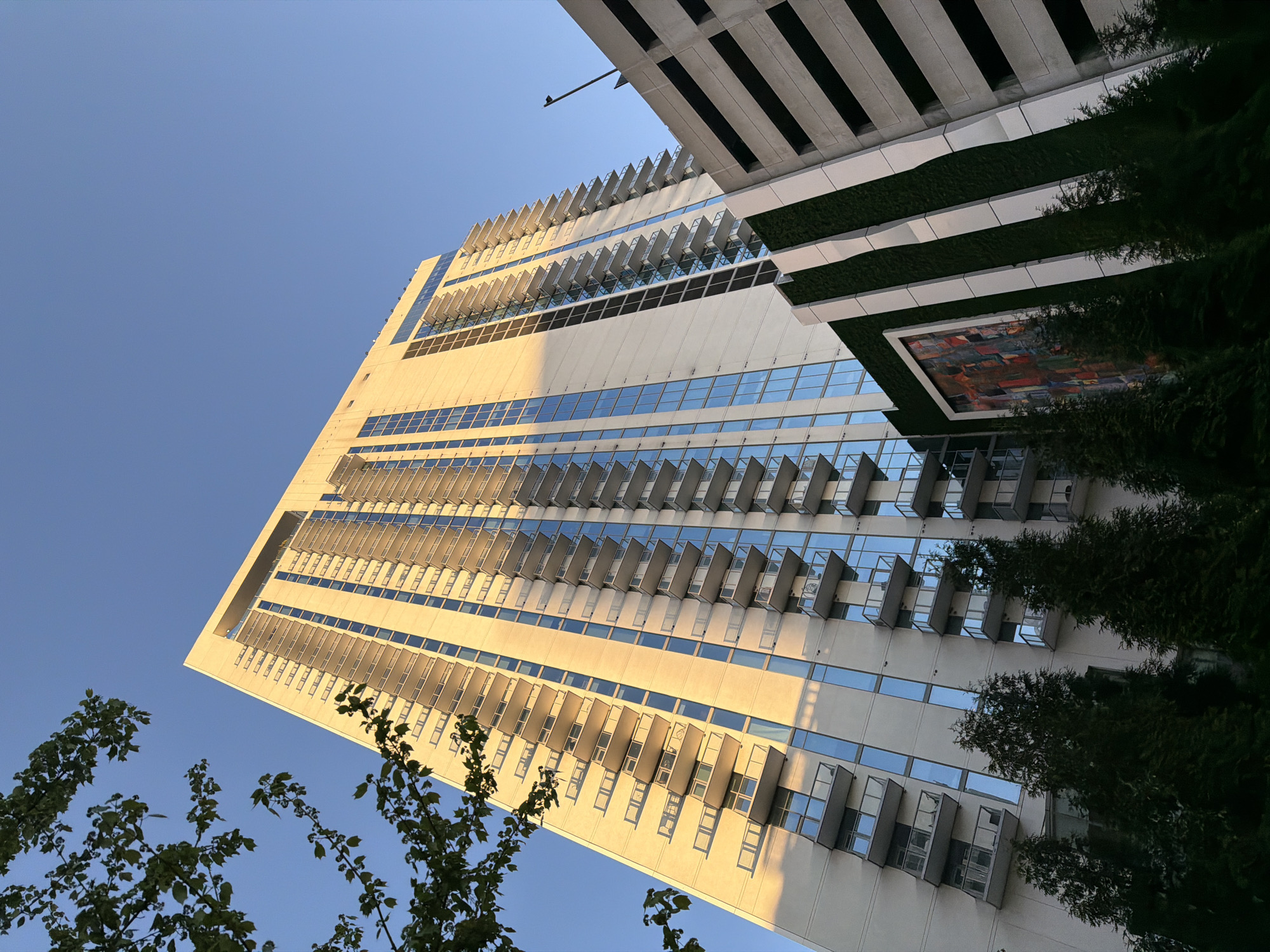 The image depicts a tall, modern building in Atlanta, USA, viewed from a low angle. The building is rectangular with a light-colored facade, and it is lined with balconies. The windows are rectangular. There is a building adjoining the first one that features white ledges. Some trees are featured in the lower portion of the image. The sky is visible in the background. The lighting suggests it is either sunrise or sunset.