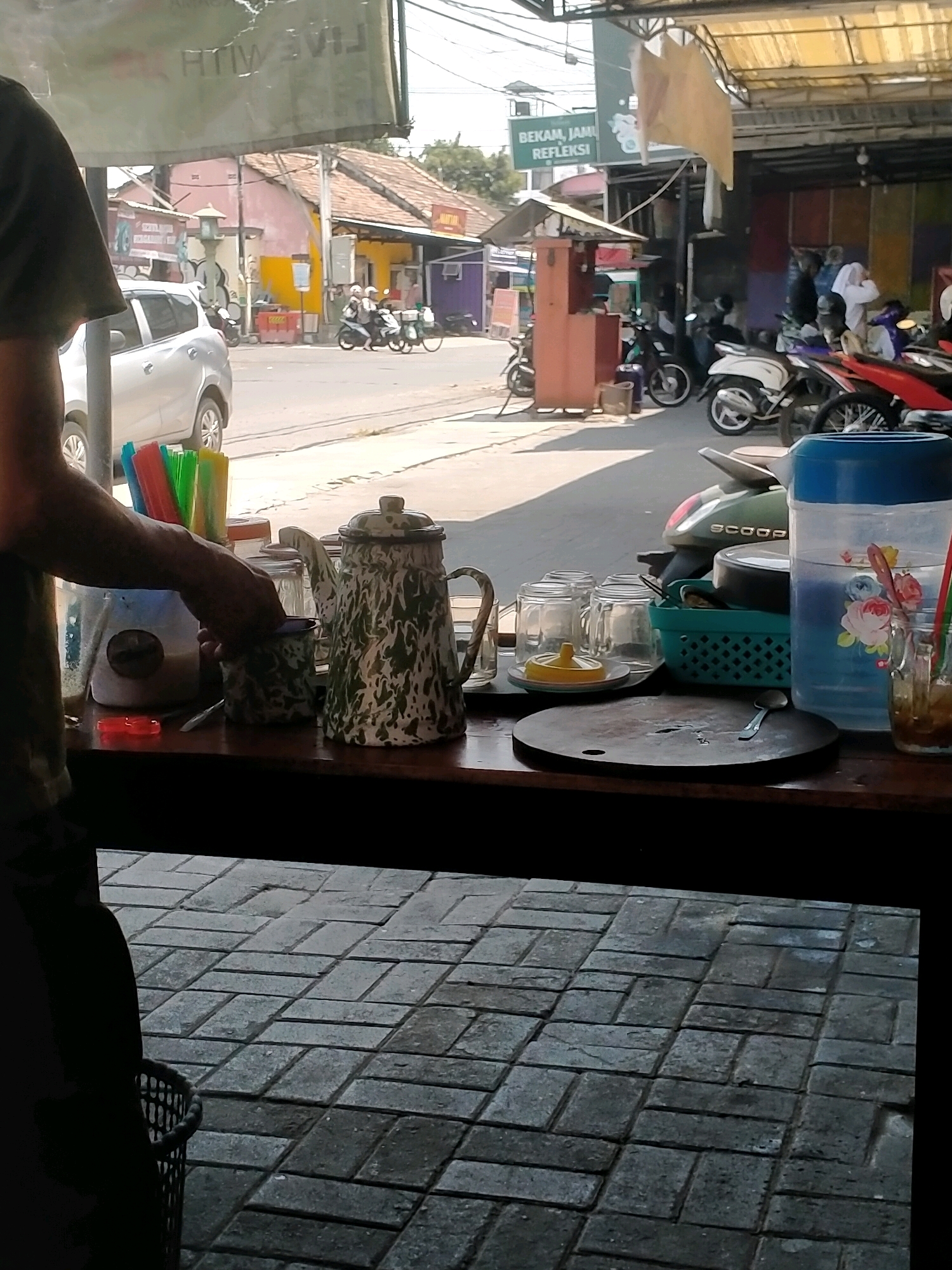 A medium shot shows a street scene outside a shop in Kasihan, Indonesia, with a table in the foreground displaying various items. The table, made of dark wood, is covered with a variety of objects including a white and green patterned enamel coffee pot, several clear glass cups, a turquoise plastic basket holding more glasses, a large blue water dispenser decorated with flowers, a cutting board with a spoon, and a clear glass with a straw and ice. A person's arm and torso are visible on the left, reaching for one of the cups. The background depicts a street with parked motorcycles and scooters, a silver car, and buildings with tiled roofs. A green sign in the distance reads "BEKAM, JAMI REFLEXI". The ground in the foreground is paved with interlocking grey bricks. The lighting suggests a daytime scene with bright sunlight.