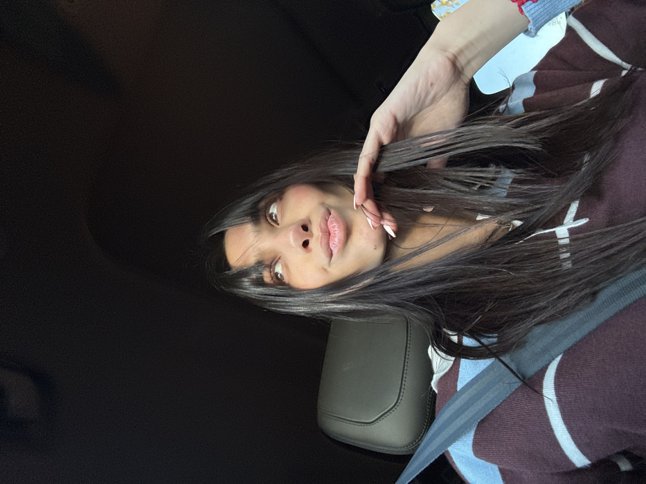 A young woman with long, dark hair and light skin is looking into the camera. She has a neutral expression. Her left hand is raised, and she is touching her hair. She appears to be sitting in a car, wearing a sweater with blue and white stripes. A seatbelt is visible across her torso. The background is dark, suggesting the car's interior.