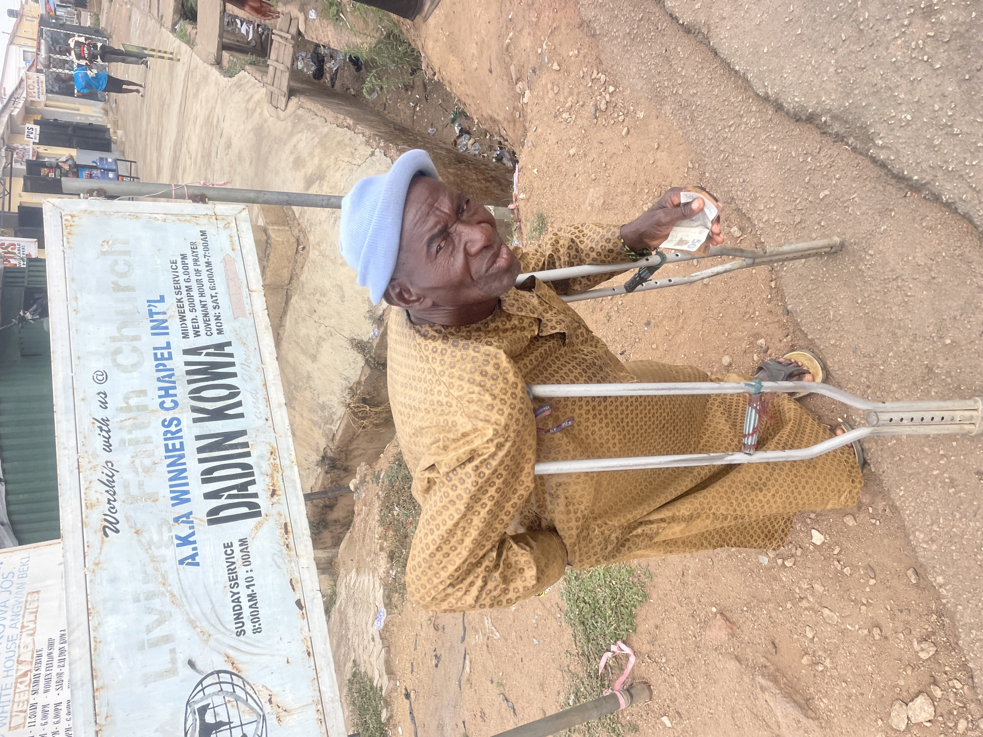 An outdoor, slightly overhead shot captures an elderly man in what appears to be a street scene in Jos, Nigeria. The man, possibly disabled, is positioned prominently in the foreground, looking directly at the camera with a serious or weary expression. He wears a light blue knitted cap, a patterned brown and gold traditional tunic, and sandals. He is supported by metal crutches, one of which he holds in his left hand, along with several folded currency notes, suggesting he may be a beggar or recently received alms.
The setting is an unpaved, dusty road mixed with some concrete sections, characteristic of a street environment in the region. To the man's left, a large, weathered white sign stands upright. This sign advertises "Worship with us @ Living Faith Church A.K.A WINNERS CHAPEL INT'L DADIN KOWA," detailing service times: "SUNDAY SERVICE 8:00AM-10:00AM," "MIDWEEK SERVICE WED. 5:00PM 6:00PM," and "COVENANT HOUR OF PRAYER MON: SAT, 6:00AM-7:00AM." Further down the street in the background, other individuals are visible, along with various buildings and smaller commercial signs, one indicating "POS AVAILABLE HERE." The ground next to the road shows some sparse vegetation and scattered debris. The bright lighting suggests it is daytime with clear weather. Another small sign partly visible in the background mentions "WHITE HOUSE ANGWAN BEKI" and "WEEKLY ACTIVITIES."