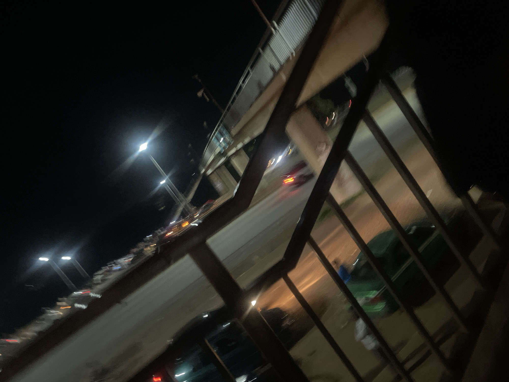 The media file shows a nighttime scene with a road and vehicles, likely in Sauka, Nigeria. The perspective is from an elevated vantage point, possibly a pedestrian bridge or balcony, looking down onto a street where cars are moving. The lighting is predominantly from streetlights and the headlights and taillights of the vehicles, creating streaks of light due to the motion and possibly a long exposure.
Several streetlights are visible, some appearing as bright, elongated glows. The road is paved, and the surrounding area is dark, indicating it's nighttime. The blurriness of the image suggests movement, either of the camera or the vehicles, or both.
On the road, there are multiple vehicles, appearing as indistinct shapes with illuminated lights. Some red taillights are visible, indicating cars moving away, and white headlights suggest cars approaching. One larger, dark green vehicle is somewhat discernible through a metal railing in the foreground.
In the foreground, dark metal railings create a visual barrier, framing the scene below. The overall impression is that of an urban or suburban environment at night, with traffic flow. The time of day is clearly night, and there are no obvious cues about the weather or specific emotions conveyed. No visible text is present in the image. The scene depicts the general activity of traffic on a road at night.