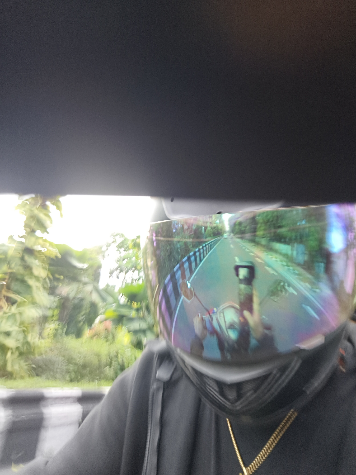 The image shows a person wearing a black hoodie and a motorcycle helmet with a reflective visor. The visor reflects a road with a white line marking, a guardrail, and lush green foliage on either side. The scene appears to be captured from the perspective of someone riding a motorcycle, looking forward and slightly down. The blurry nature of the image suggests motion. The presence of lush greenery and the style of the guardrail are consistent with tropical environments. While the exact location cannot be definitively determined from the image alone, the mention of Denpasar, Indonesia, aligns with the visual cues of a tropical, possibly urban or suburban, setting with a paved road and abundant vegetation.