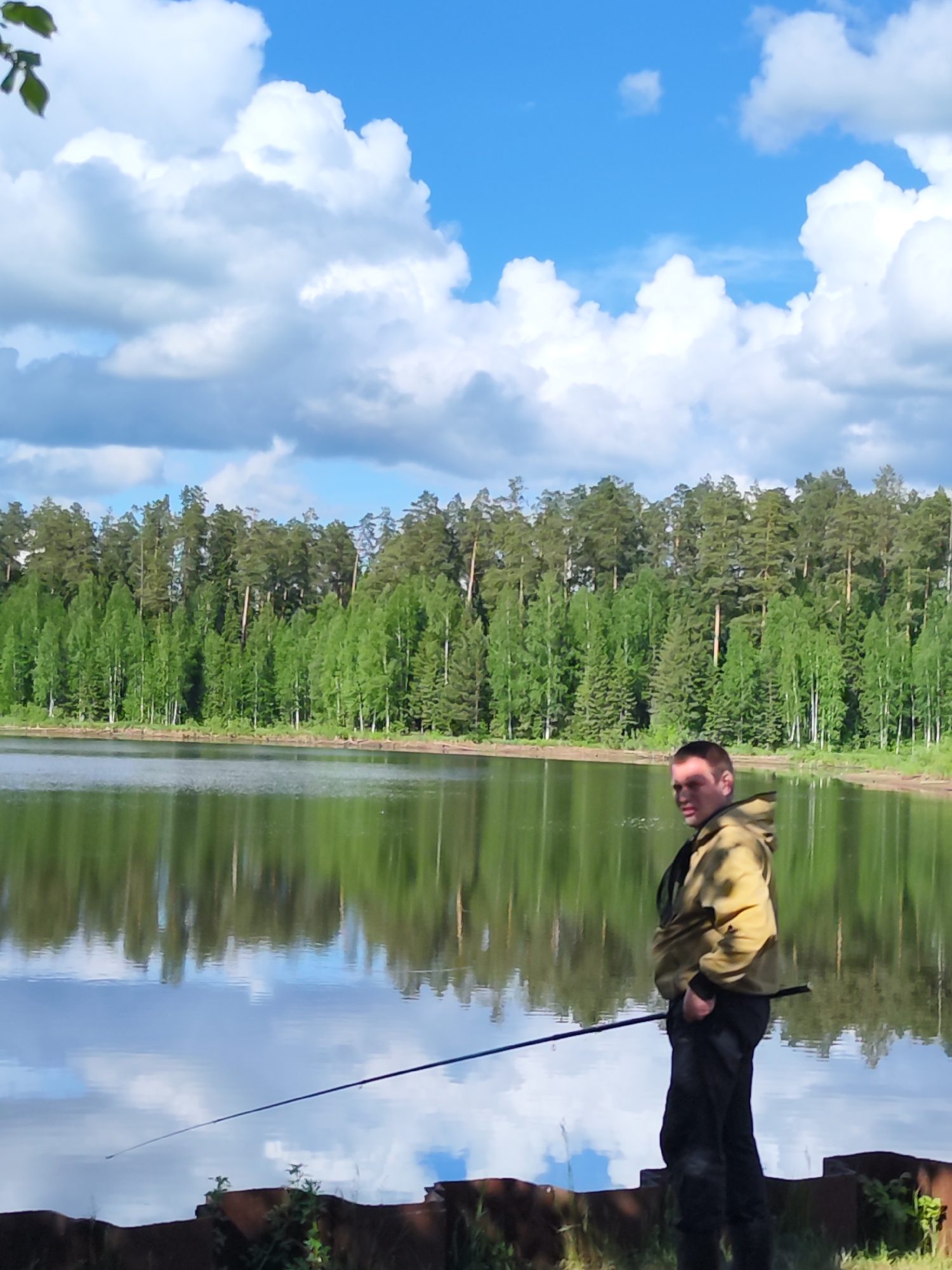 A man stands on a bank fishing in Rusalevka, Russia. The body of water reflects the sky and the trees lining the opposite shore. He is wearing dark pants, a yellow jacket, and is holding a fishing rod. The sky is blue with white clouds. The scene is outdoors in a natural setting.